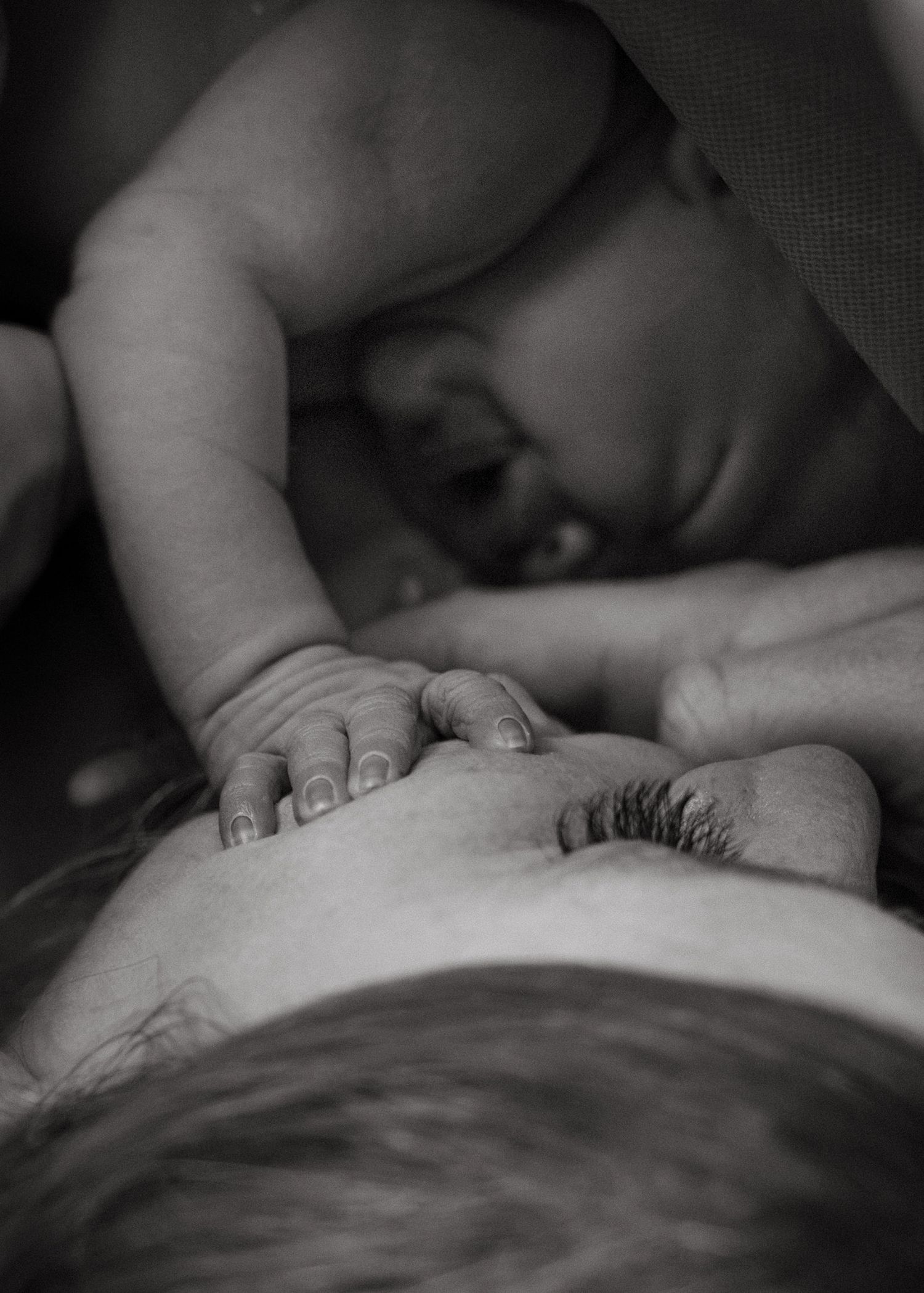 Black and white photo of a newborn's small hand touching a mother's face as she rests her eyes closed beside her baby.