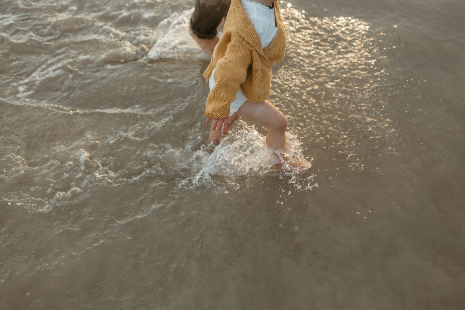 Toddler in a yellow cardigan splashes through shallow shimmering ocean waves along the shoreline.
