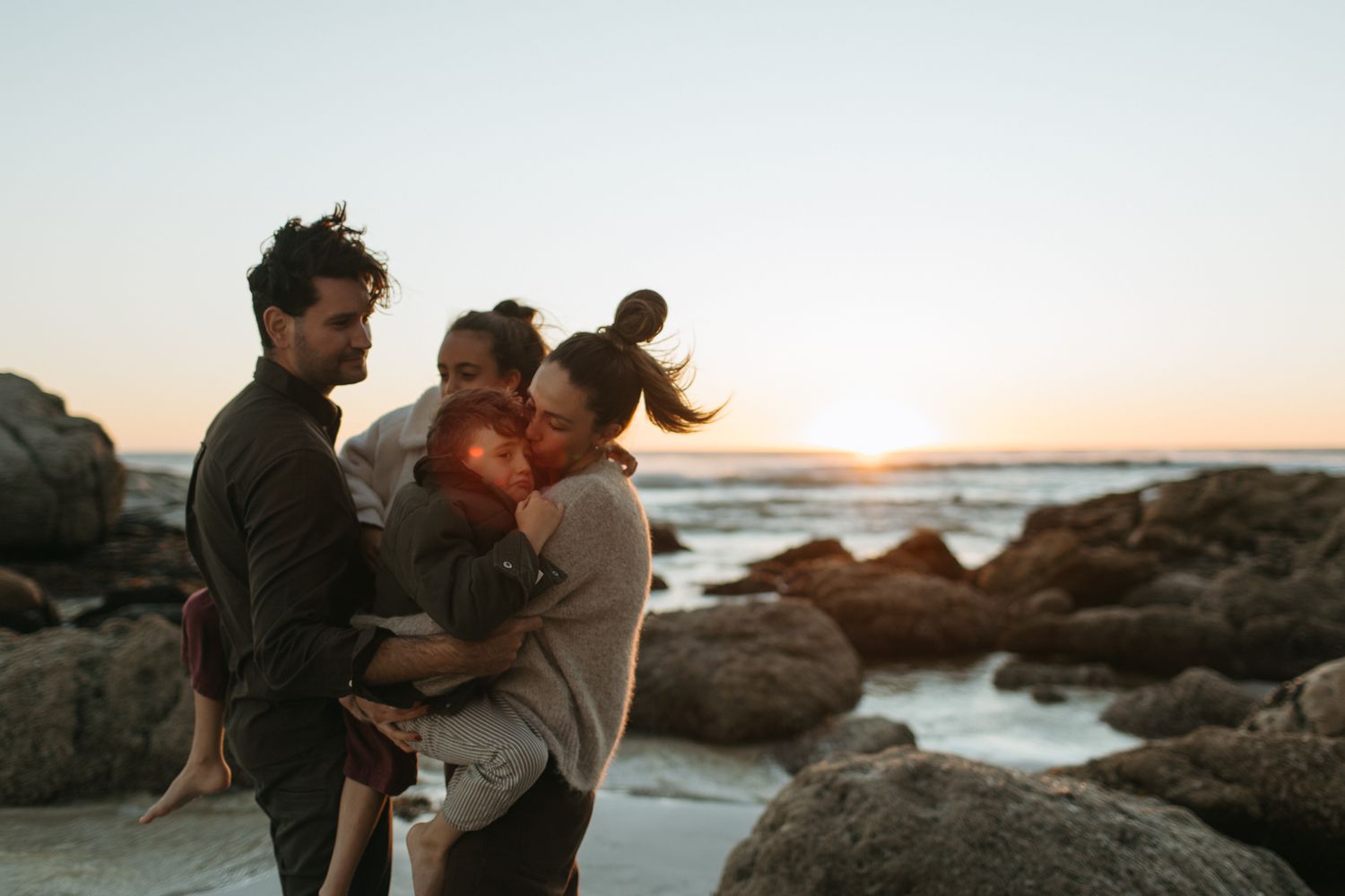 A family of four embraces on a rocky beach at sunset, with the parents carrying their two young children.