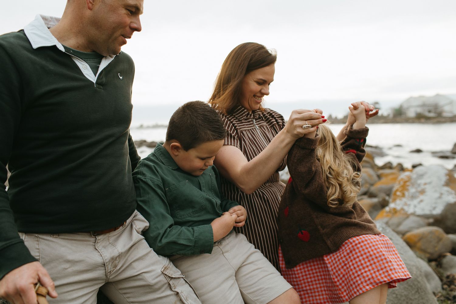 A smiling family of four sits together on rocky shoreline, with a boy in green sweater and a girl in plaid skirt beside their parents.