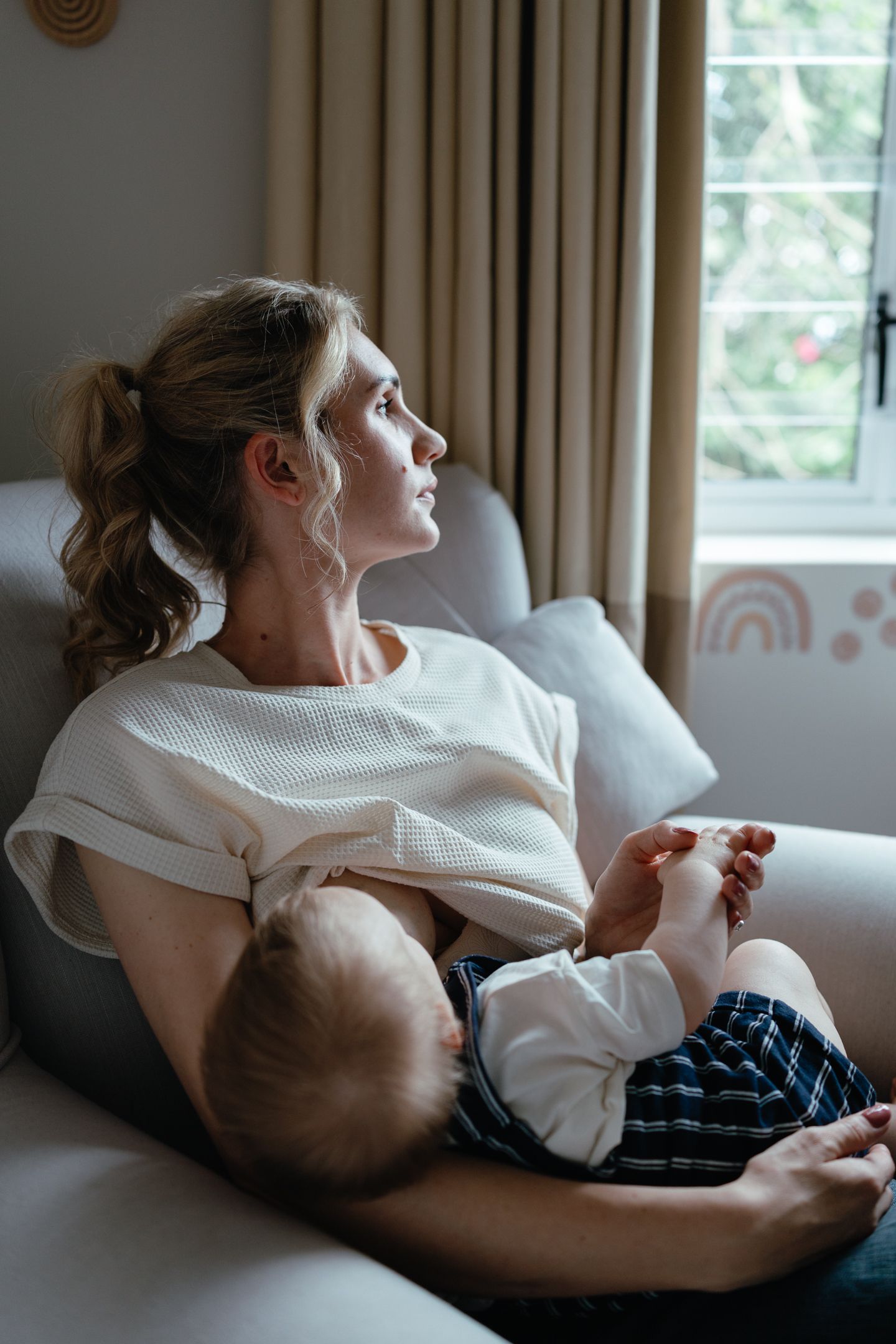 Mother with a ponytail looks out a window while holding her baby in a nursery with a rainbow decal.