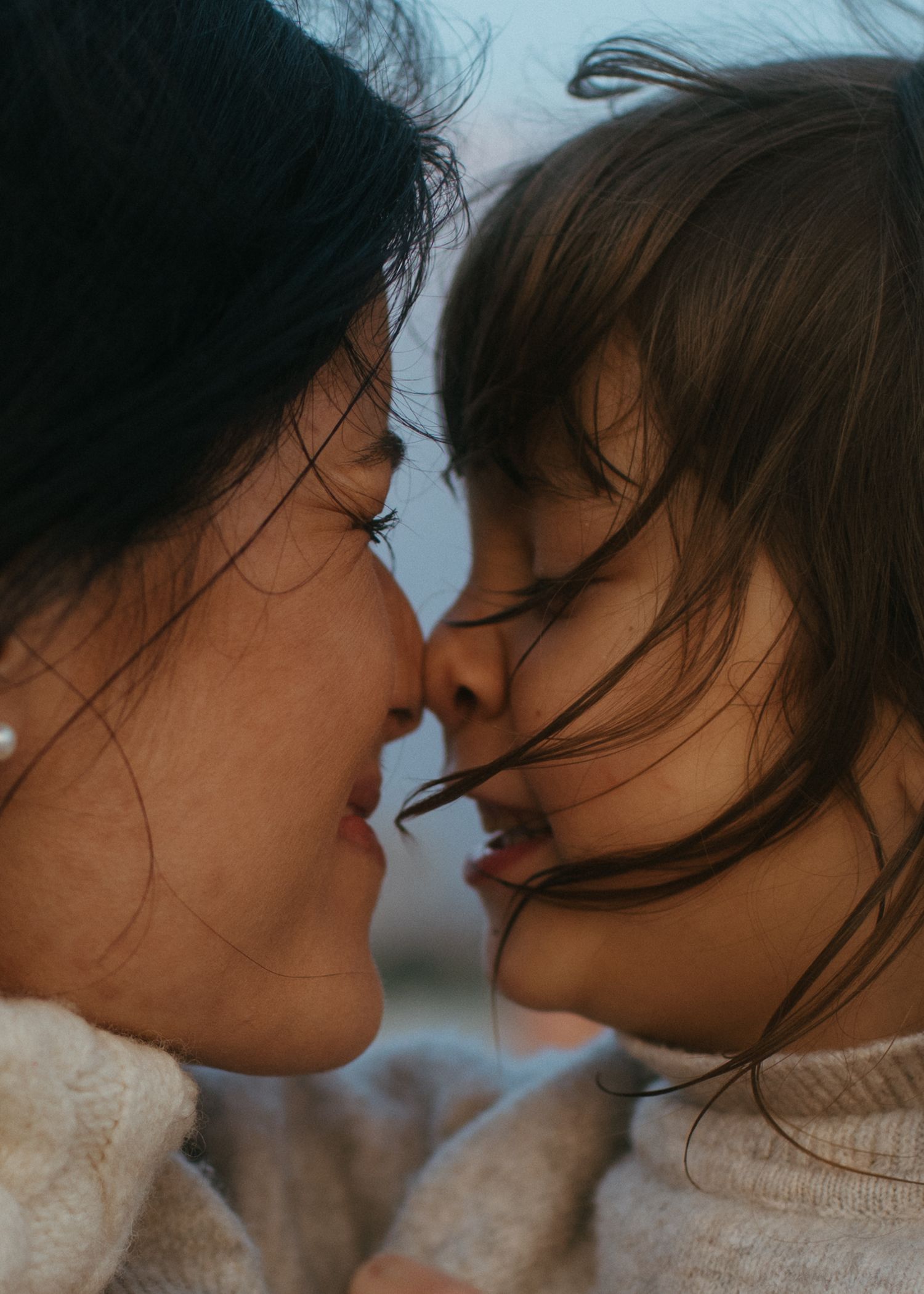 Close-up of a mother and young child touching noses affectionately with windblown hair at the beach.