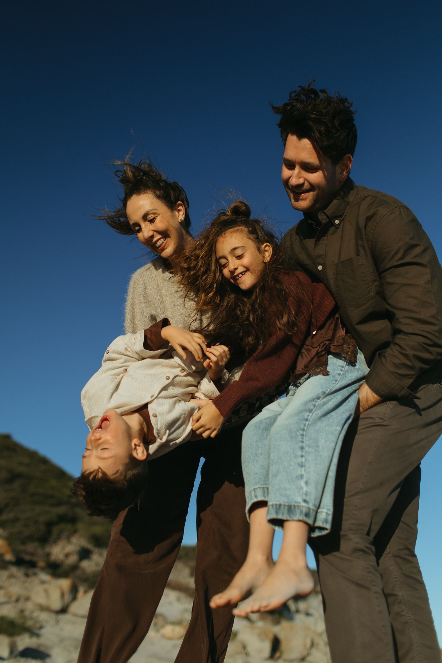 A smiling family plays on a rocky coast under a clear blue sky as parents swing their kids joyfully.