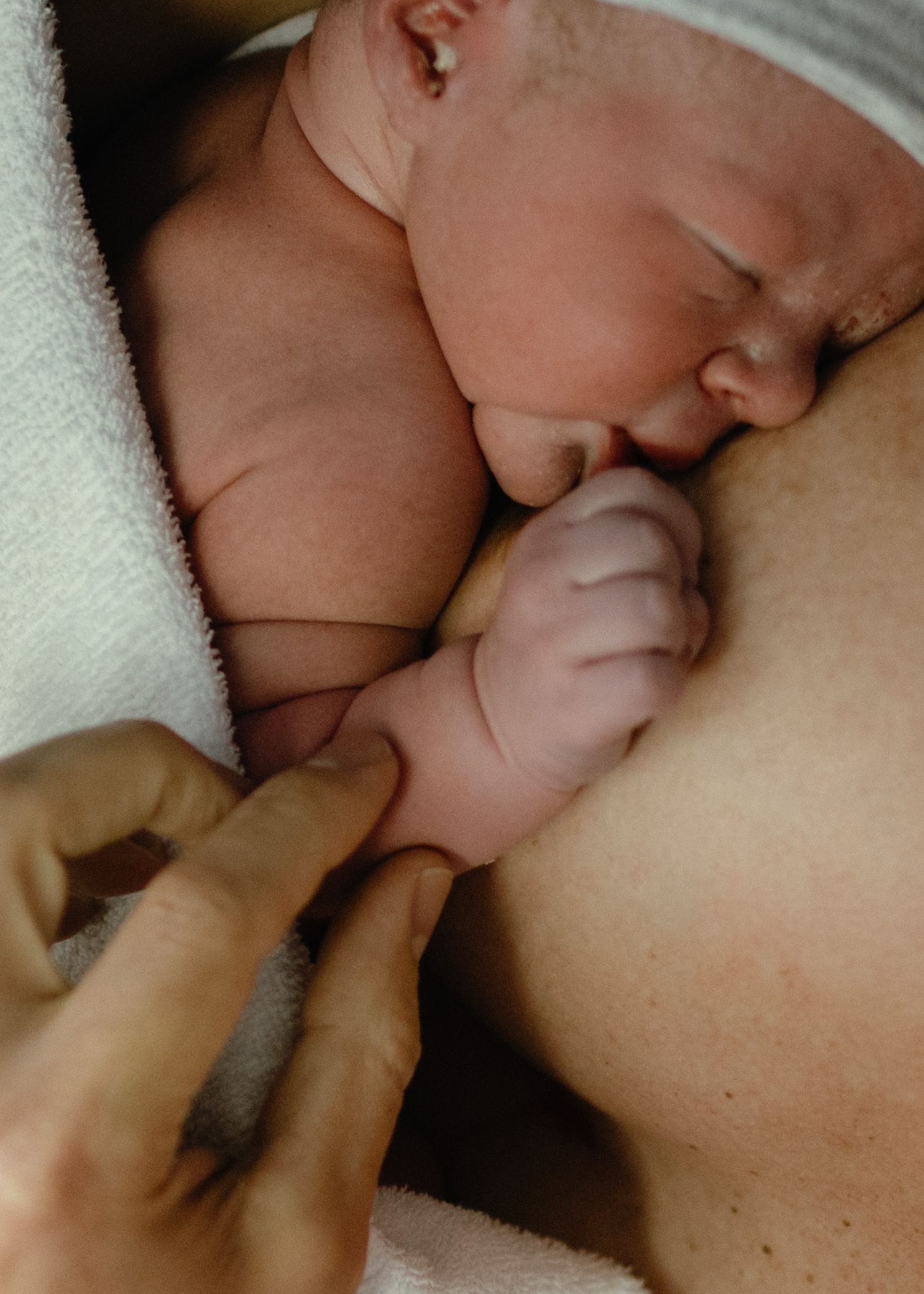 Close-up of a newborn baby nursing at its mother's chest, tiny hand resting on skin, while an adult hand gently touches the baby's arm.