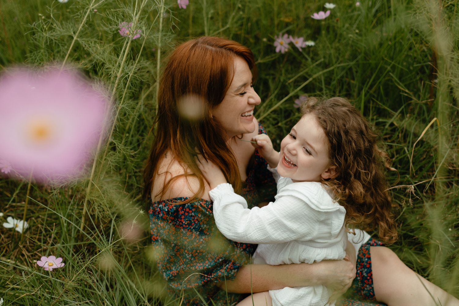 Red-haired mother and her giggling daughter cuddle together in a grassy meadow with pink wildflowers.