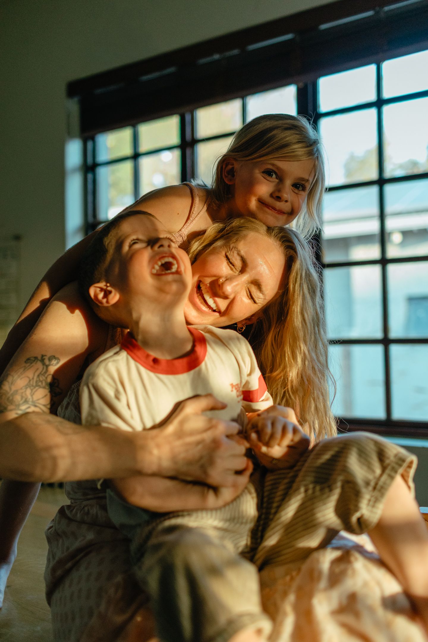 A tattooed mother laughs with two happy children in a sunlit room beside a large window.
