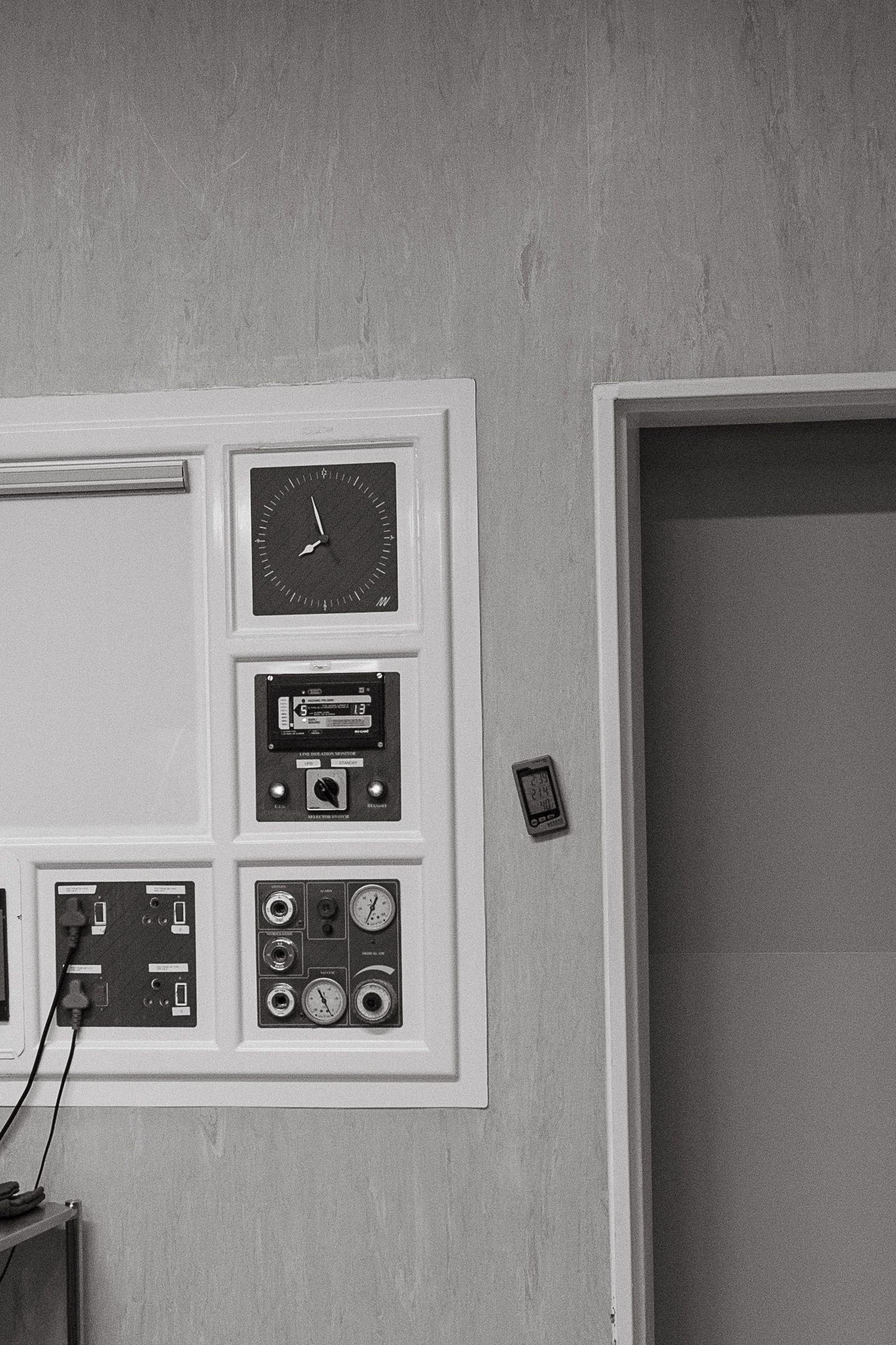 Black and white photo of a wall-mounted control panel with a clock, gauges, switches and cables beside an open doorway.