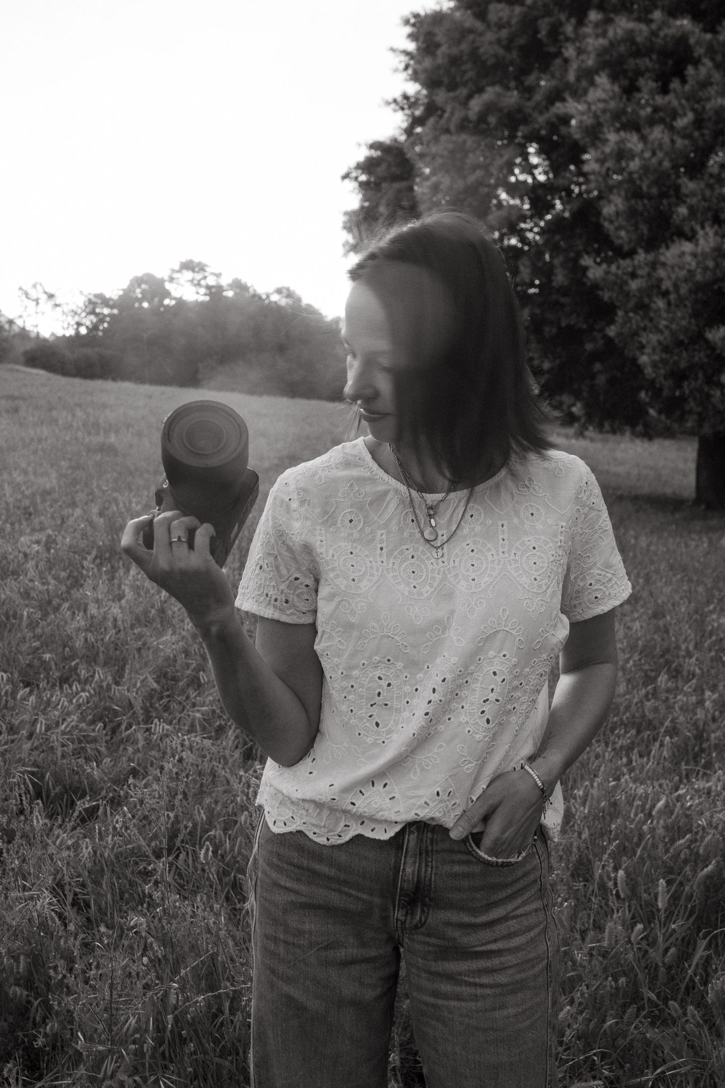A woman in a white eyelet blouse and jeans looks down at a camera lens in her hand while standing in a grassy field.