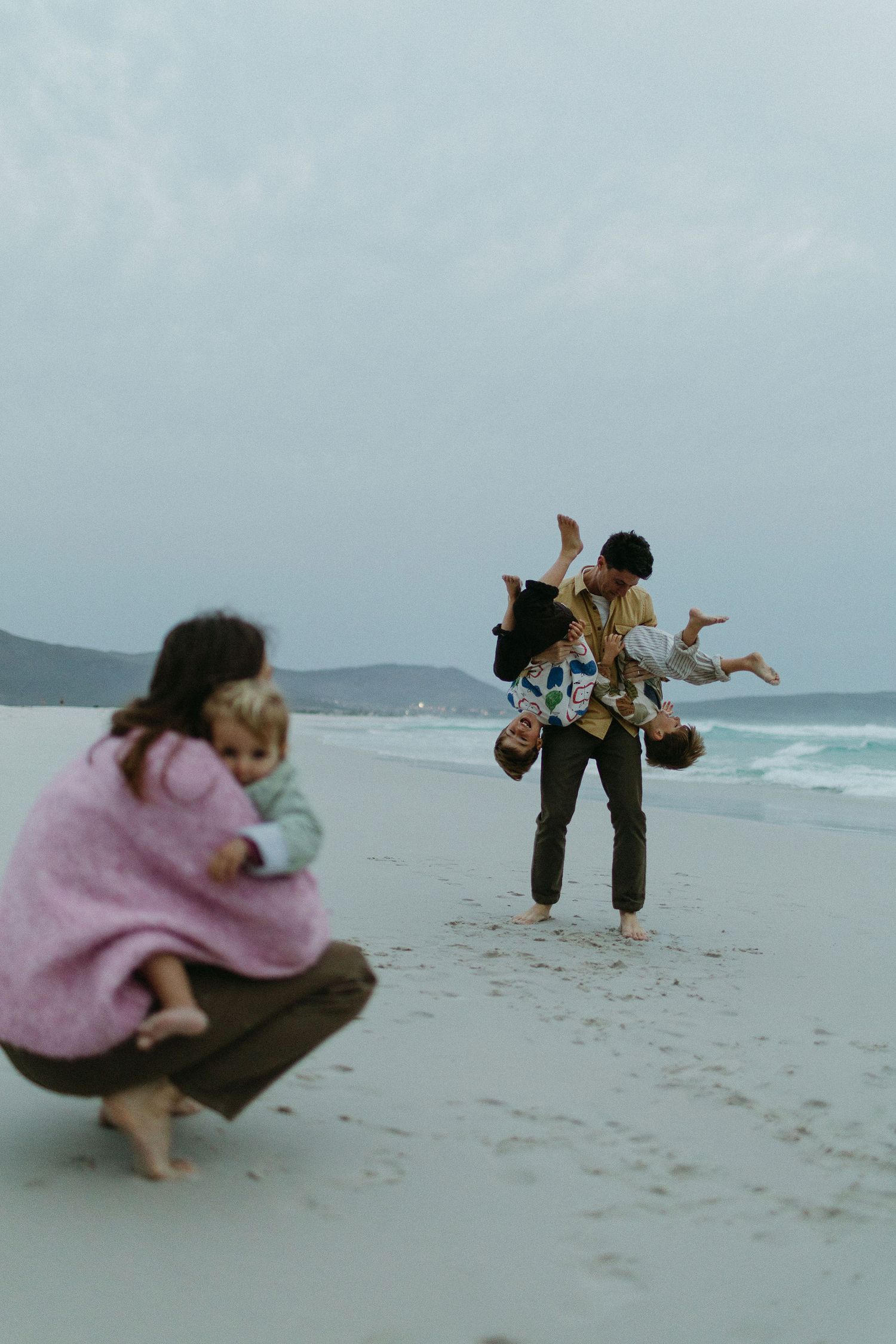 A father swings a child upside down on a quiet beach while a mother crouches nearby holding a toddler.