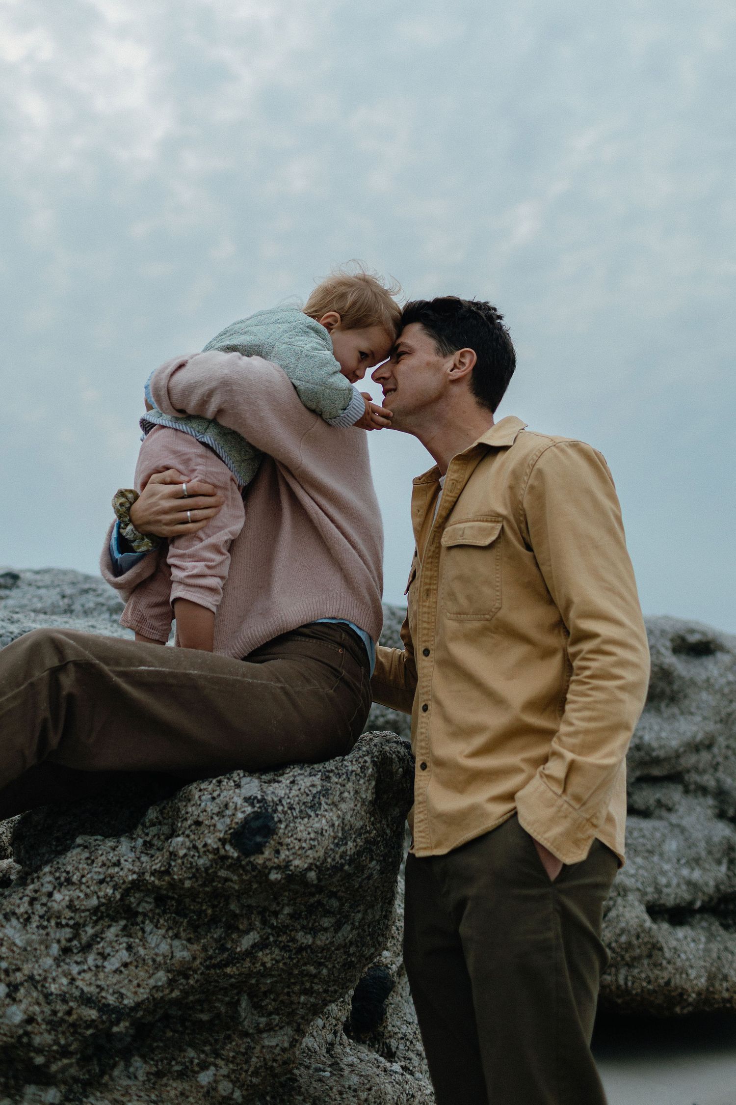 A father touches foreheads with his toddler, held by the mother, on a rocky outcrop under a soft cloudy sky.