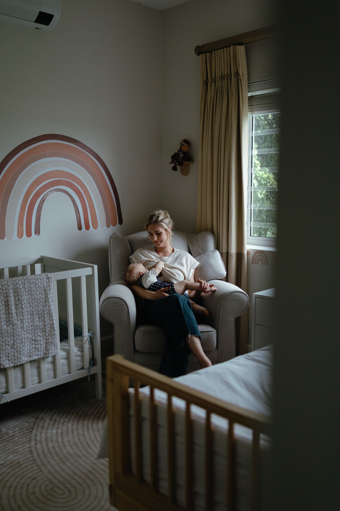 A mother sits in a gray armchair holding her baby in a nursery with a rainbow wall decal, white crib and sunlit window.