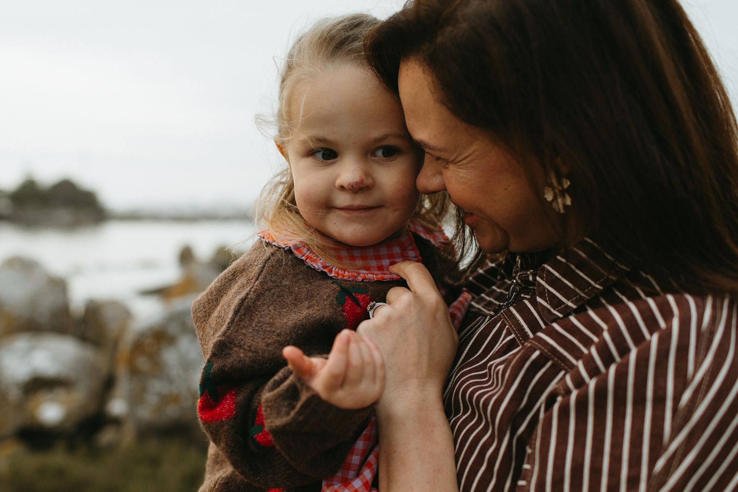A smiling mother holds her curious young daughter close beside a rocky coastline, sharing a warm and affectionate family moment.