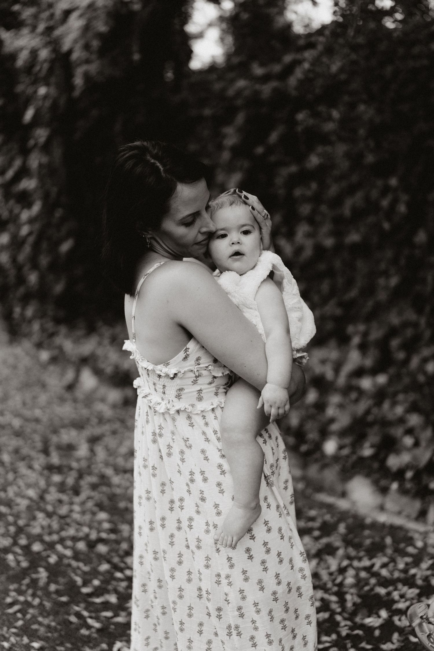 A black and white image of a mother tenderly embracing her smiling baby girl in a leafy outdoor setting.
