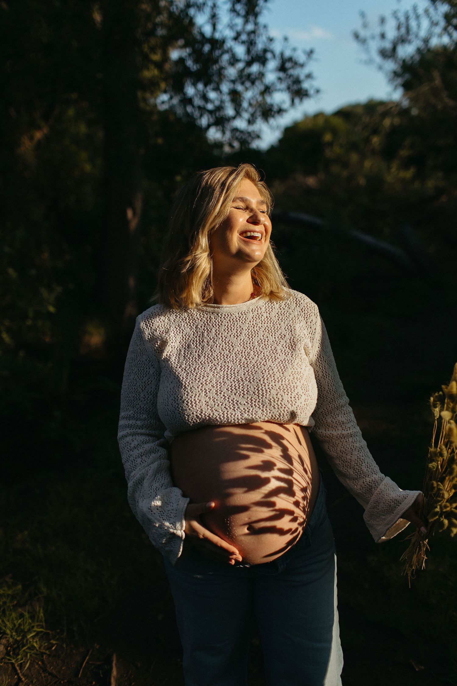Laughing pregnant woman in a knit sweater stands in golden sunlight as leaf shadows dapple her bare belly outdoors.