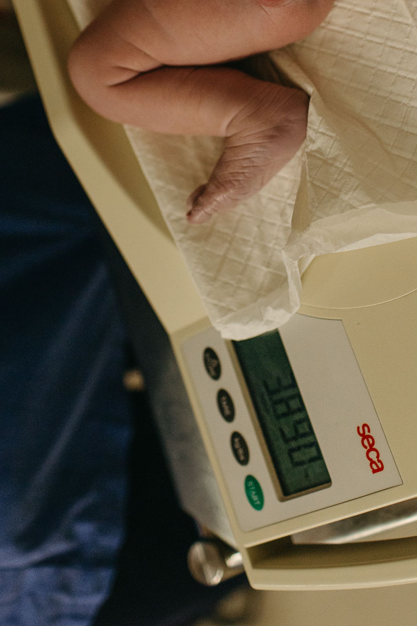 Newborn baby's tiny foot resting on a white paper-lined Seca digital scale during a weigh-in, with the display visible below.