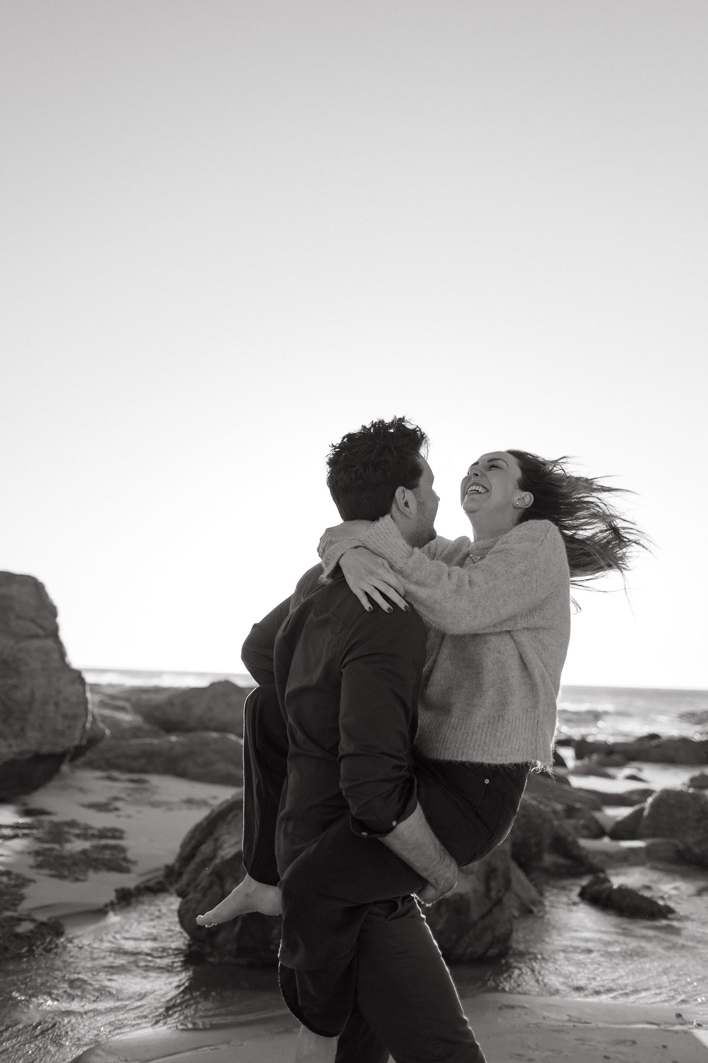 Black and white photo of a man lifting a laughing woman in his arms on a rocky beach at sunset.