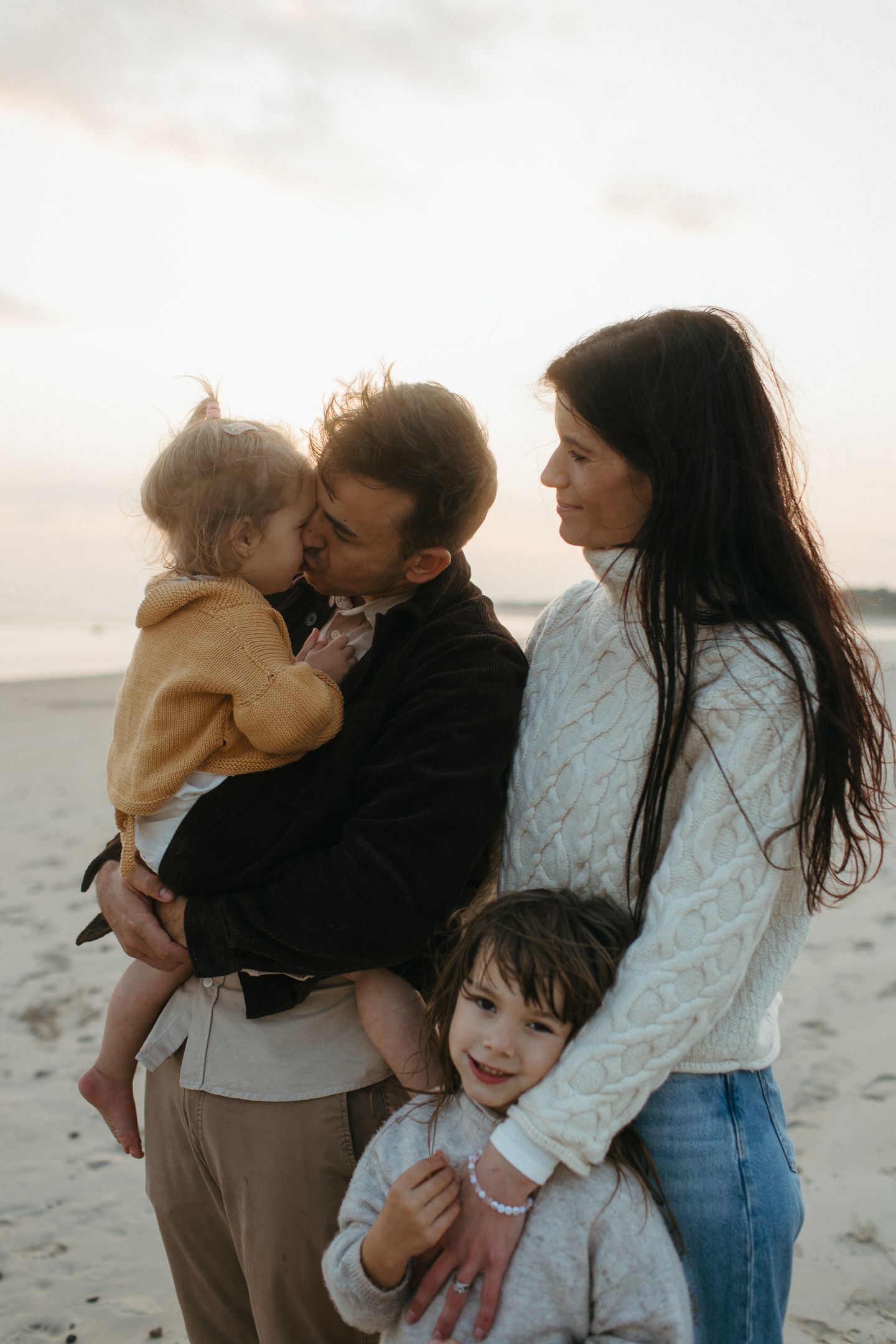 Family of four embracing on a beach at sunset as a father kisses his toddler and a mother hugs her daughter.