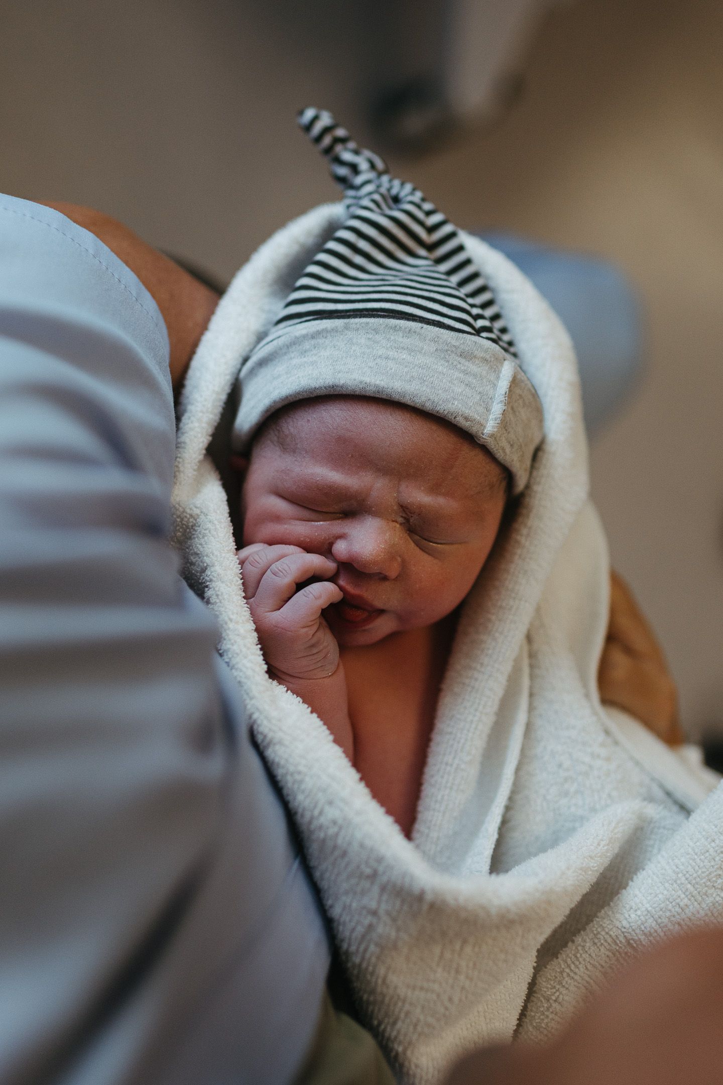 Newborn baby in a striped knotted hat wrapped in a soft white towel scrunches its face while held in a parent's arms.