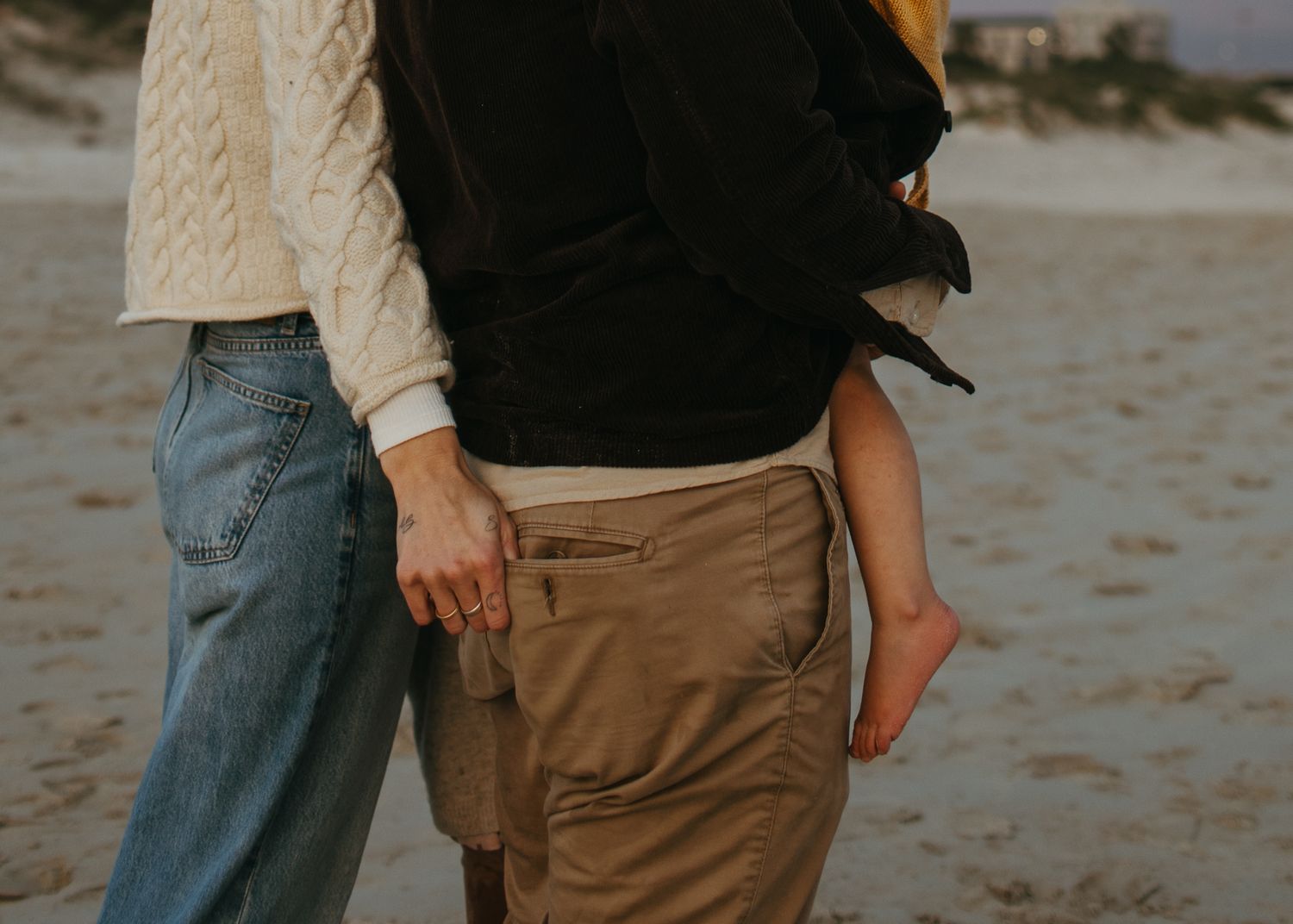 Parents standing close together on a sandy beach, holding a toddler whose bare foot dangles at their side.