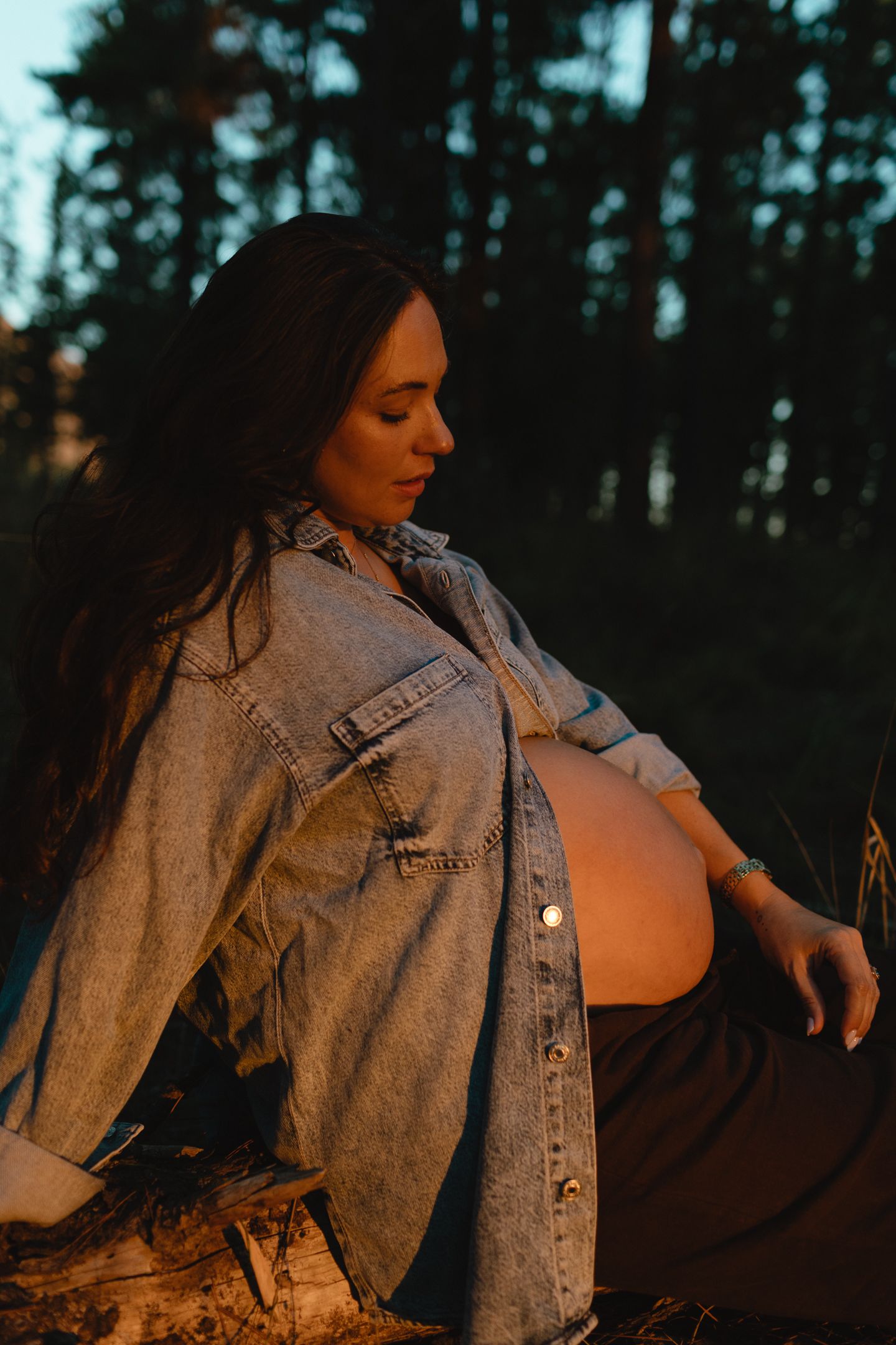 A pregnant woman in a denim jacket sits on a log in a forest, her profile lit by warm golden sunset light.