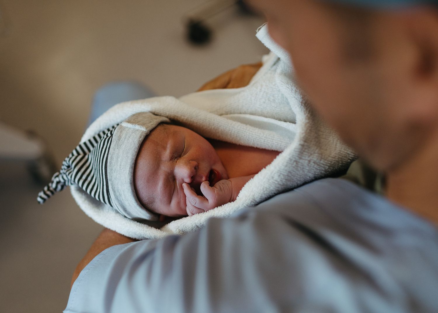 Newborn baby wrapped in a white blanket and striped hat sleeps while being held in a hospital.