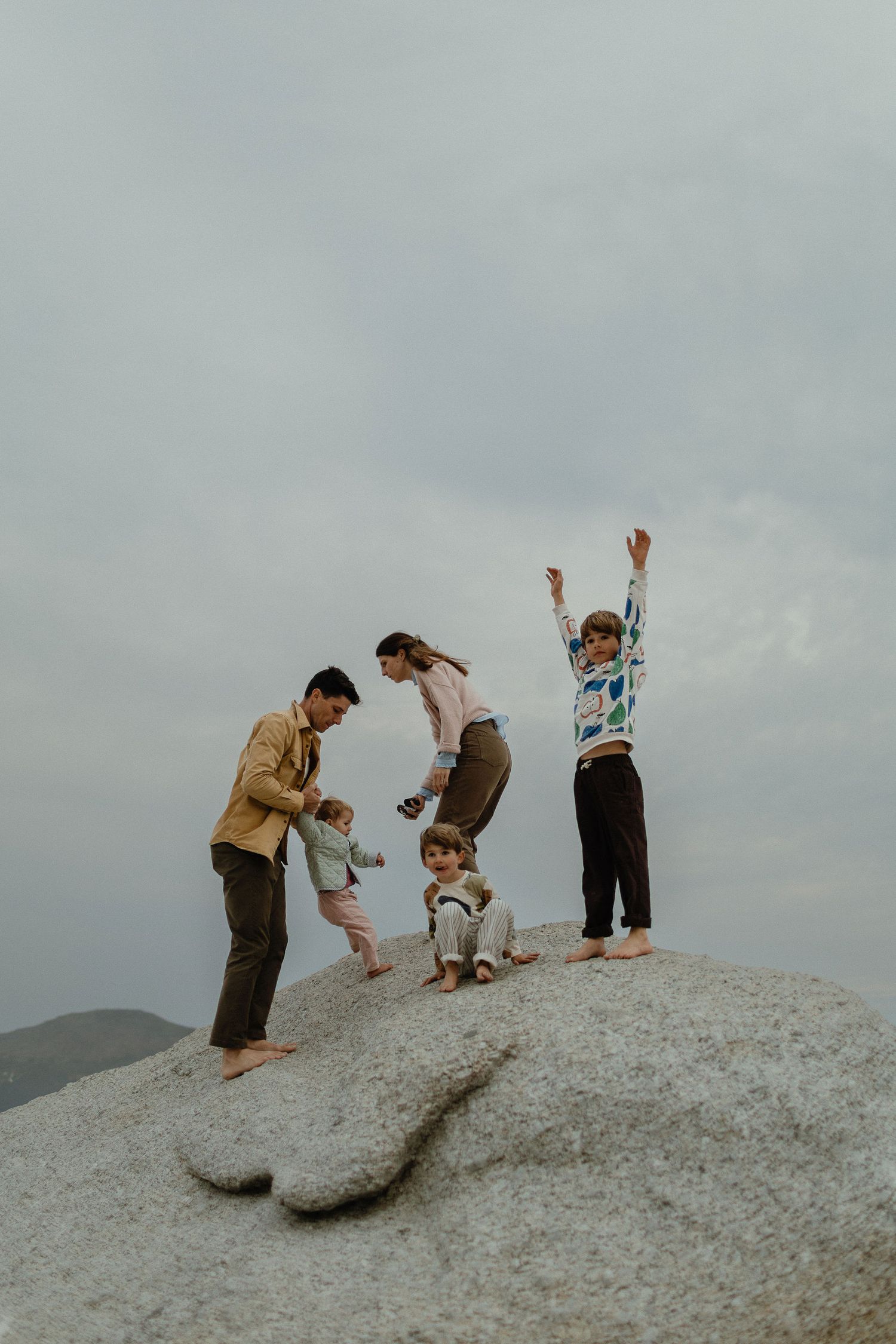 Family of five stands on a large granite boulder under a gray sky, with a child raising both arms in the air.