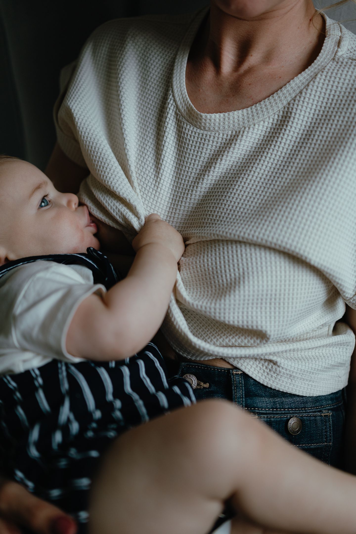 Close-up of a baby in a striped outfit nursing and reaching for the cream waffle-knit top of a mother in an intimate breastfeeding moment.