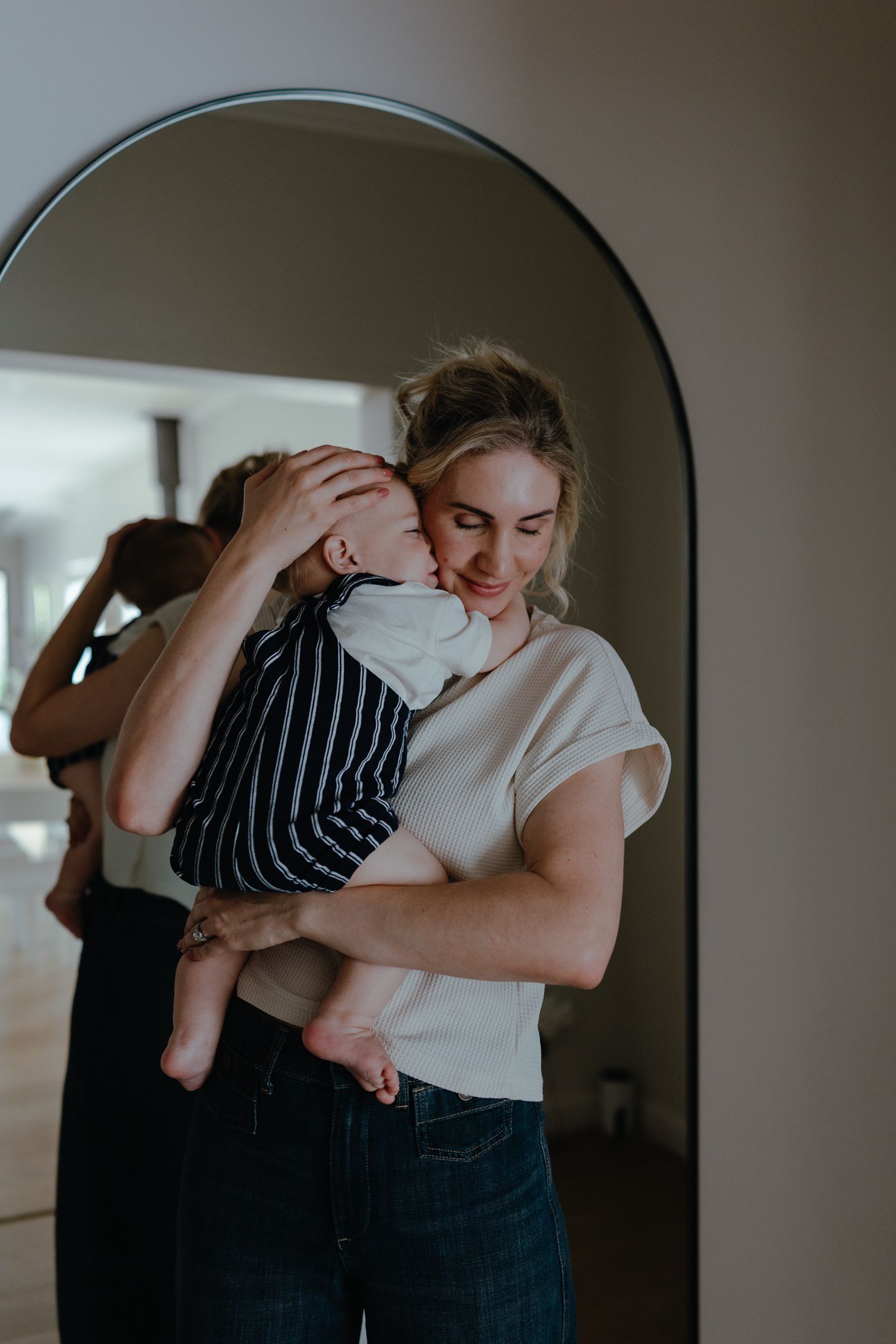 Smiling mother in a cream top cuddles her baby in front of an arched mirror during a relaxed lifestyle newborn photo session at home.