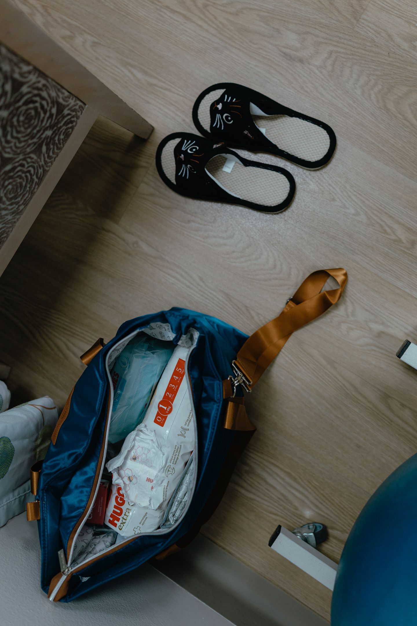 Open blue and tan duffel bag packed with Huggies diapers and wipes beside black cat-face slippers on a light wood floor.