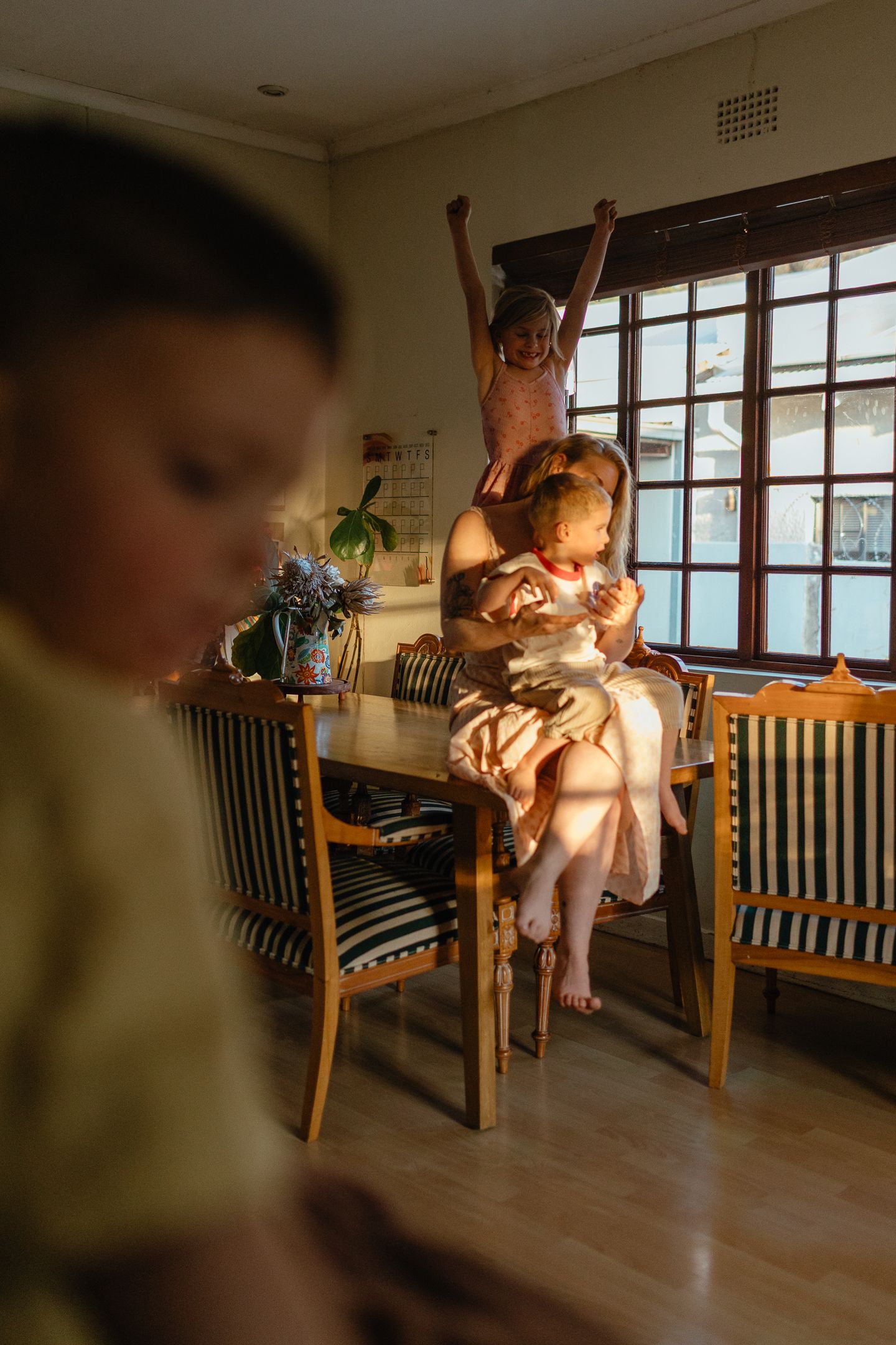 A mother holds a toddler at a wooden dining table in warm window light while a girl behind them cheers with raised arms.