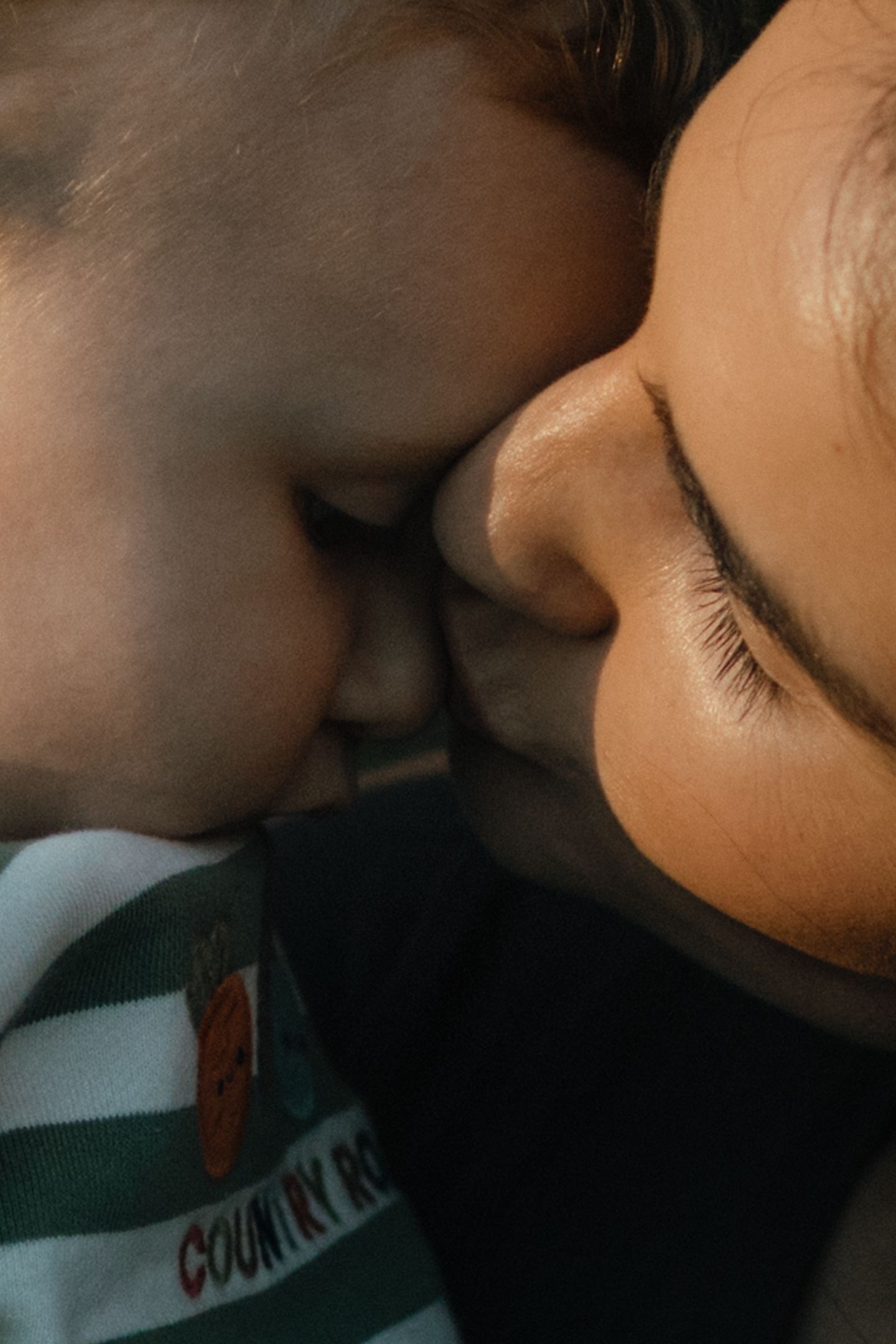 Close-up of a mother gently kissing her baby's cheek, the child wearing a green striped outfit with a carrot patch.