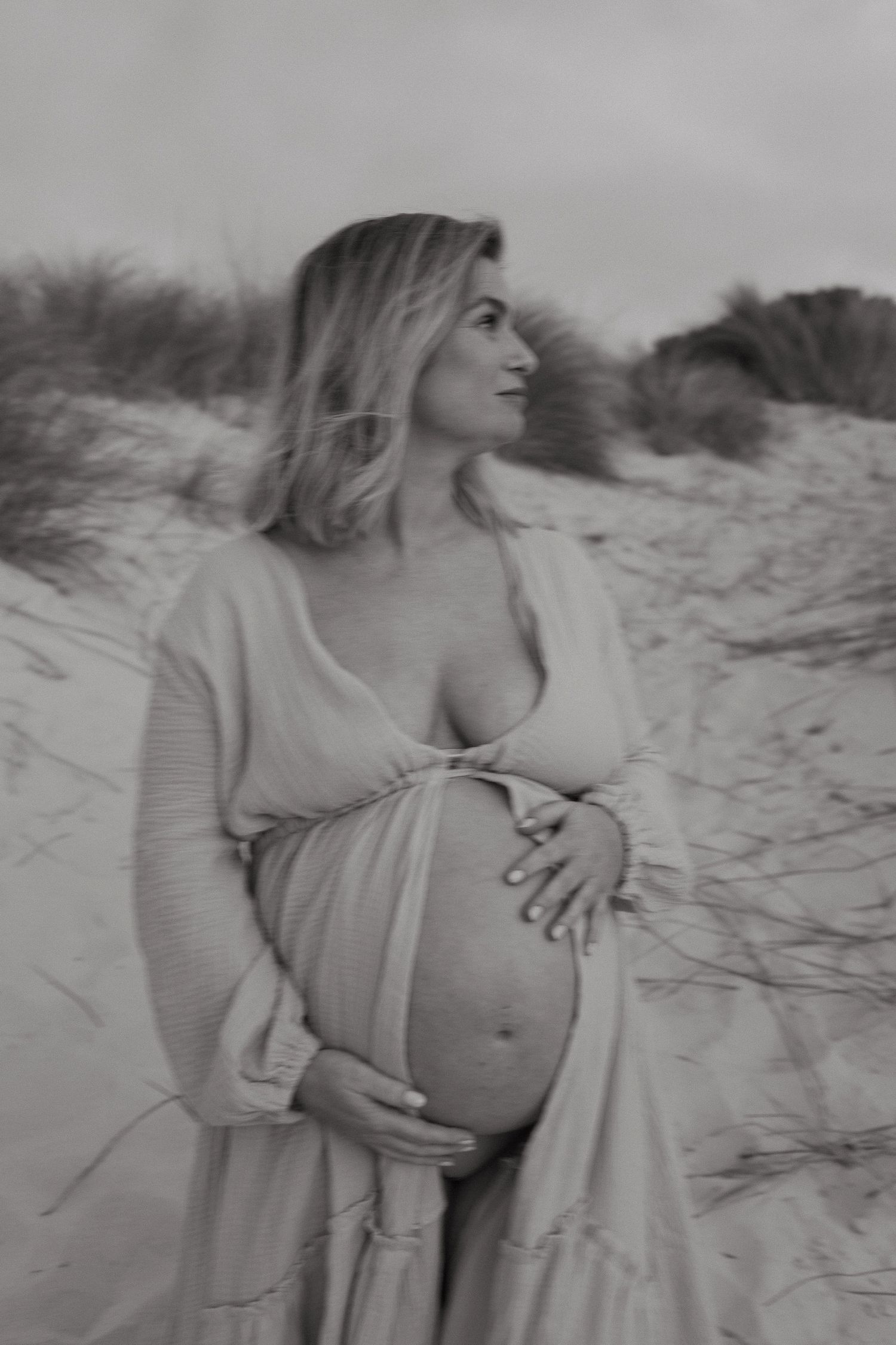 Black and white maternity portrait of a pregnant woman in a flowing robe cradling her belly on a sandy beach dune.