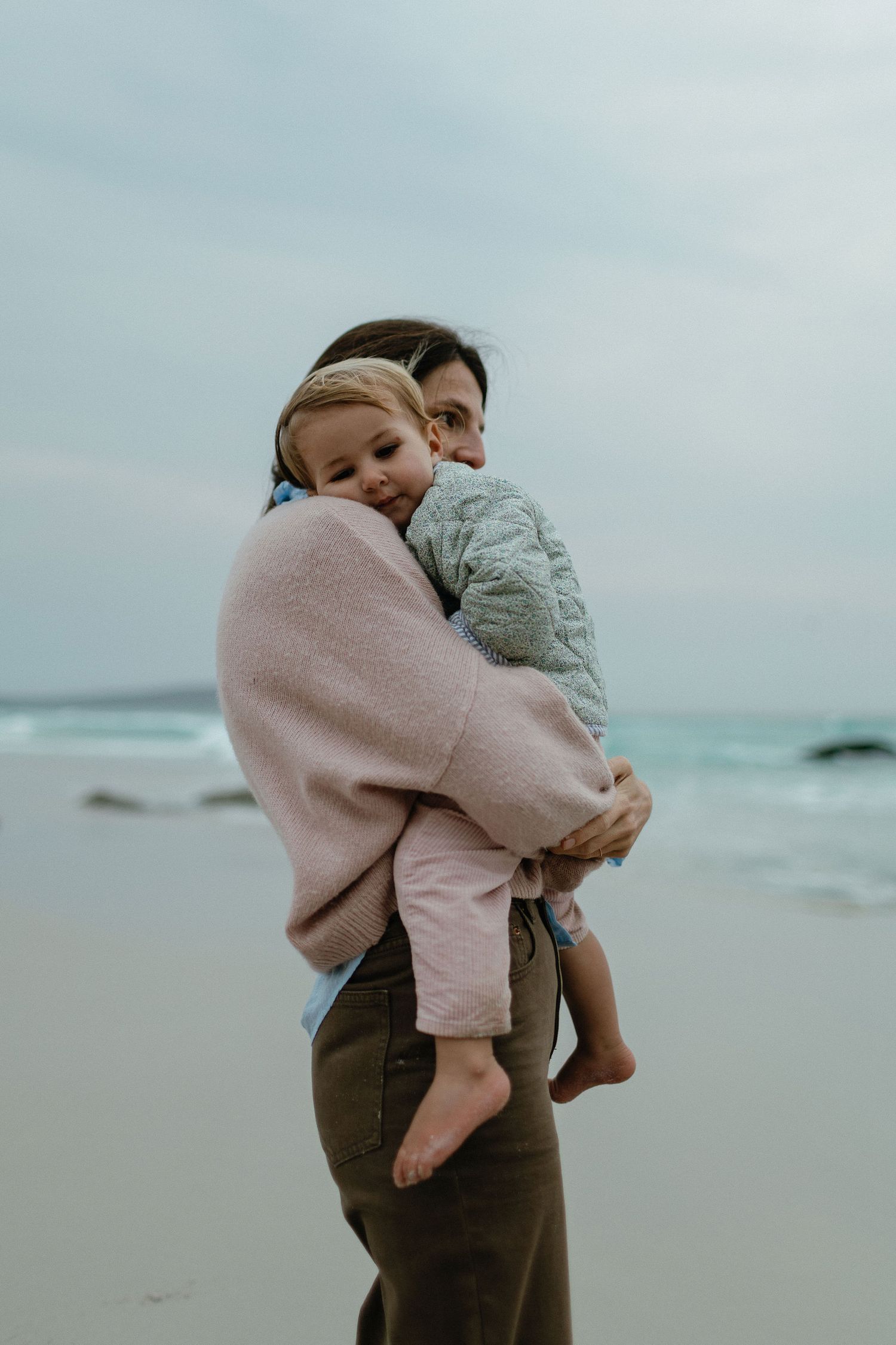 A mother in a pink sweater carries a sleepy toddler in her arms on a quiet beach under a soft overcast sky.