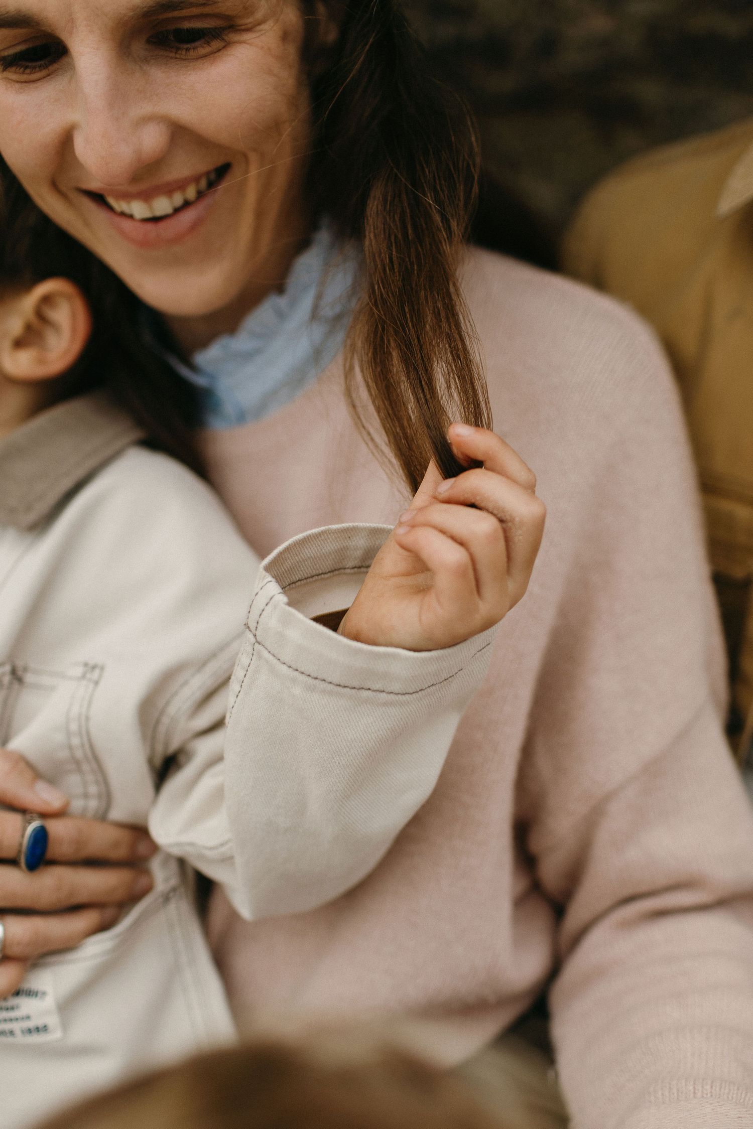Smiling woman in a soft pink sweater looks down at a small child in a cream jacket during a warm, natural family portrait.