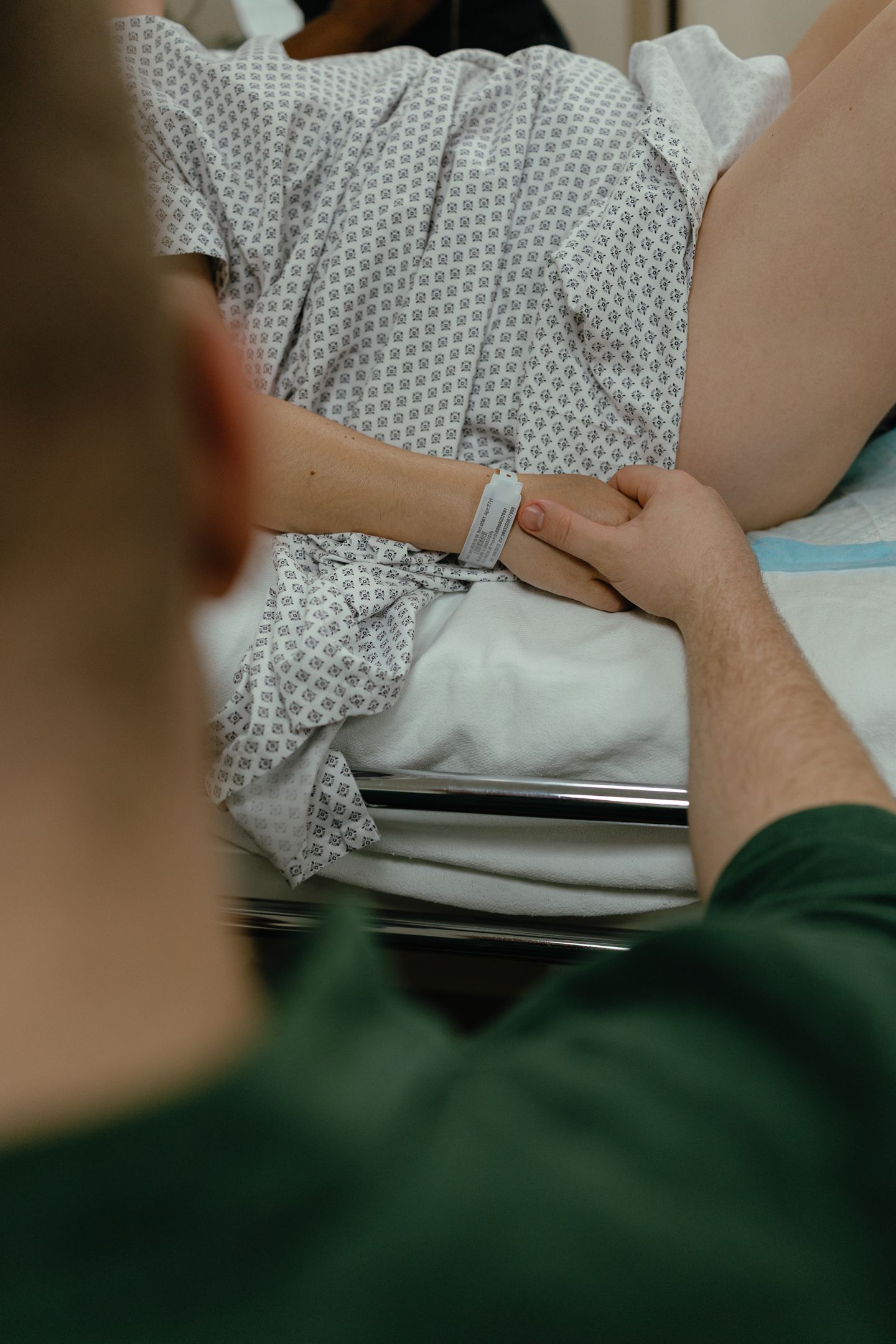 Partner in a green shirt holds the hand of a patient in a hospital gown wearing an ID wristband while she lies on a hospital bed.