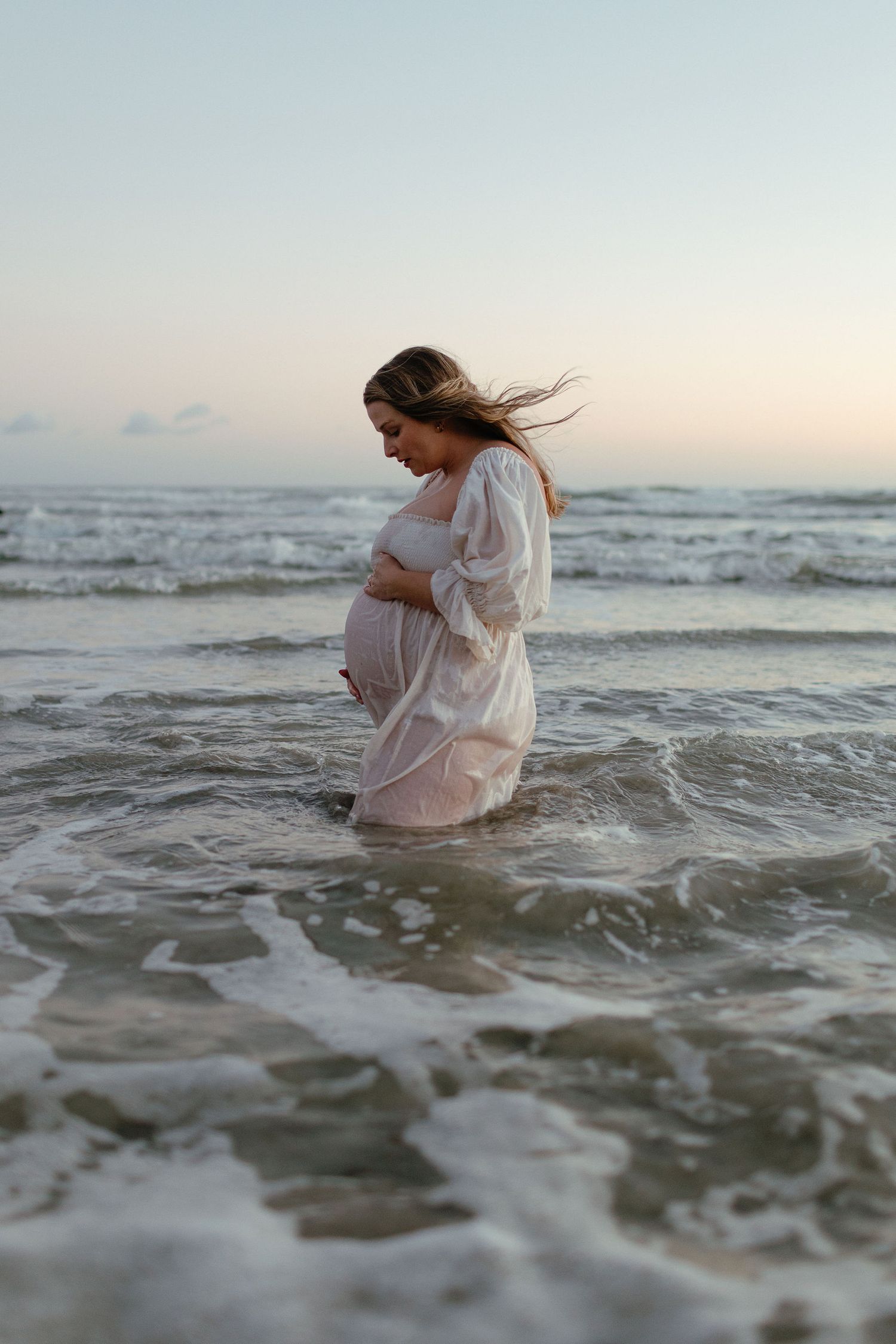 Pregnant woman in a flowing white dress cradles her belly while standing in ocean waves at sunset.