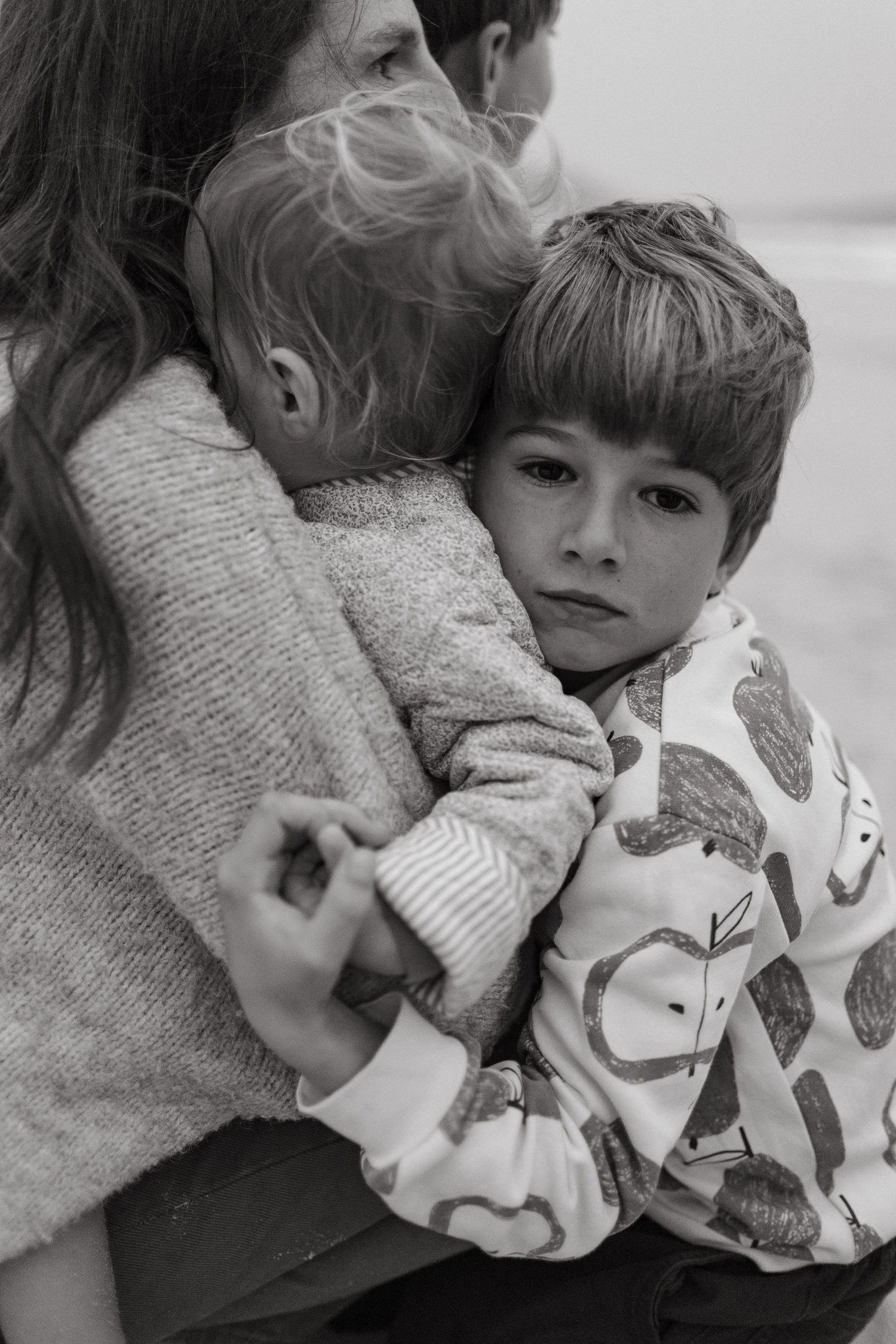 Black and white portrait of a thoughtful boy in an apple-print shirt hugging his mother and toddler sibling.