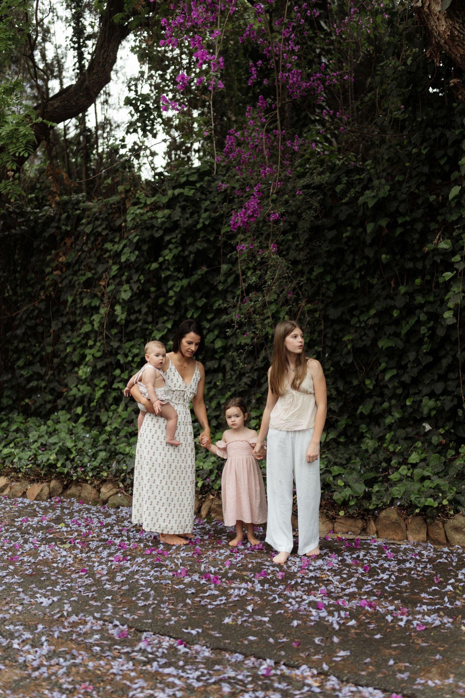 A mother holding her baby stands beside two daughters on a path covered in purple petals beneath a blooming tree.
