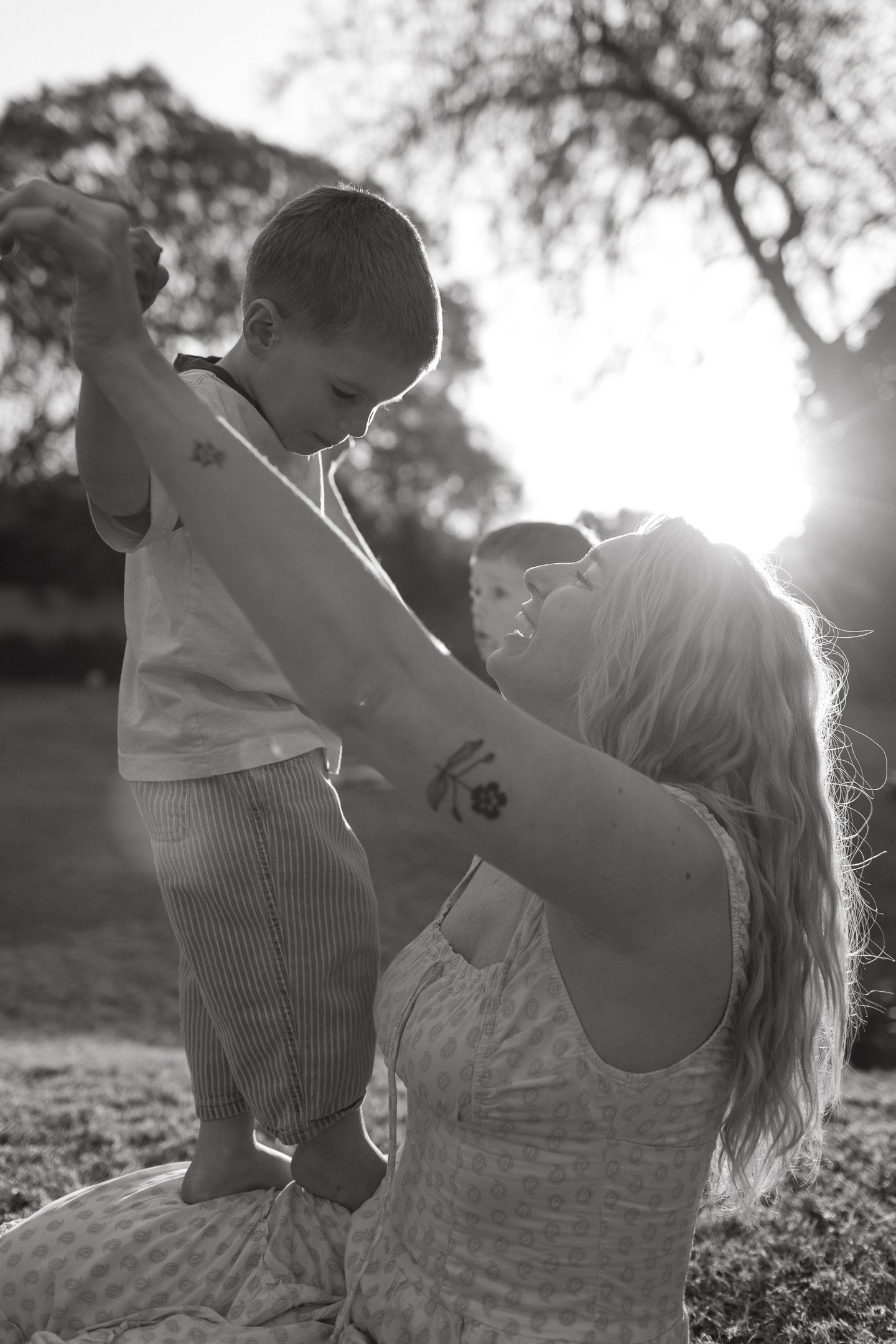 Black and white photo of a smiling mother lifting a toddler's hands as he stands on her lap in a sunlit park.