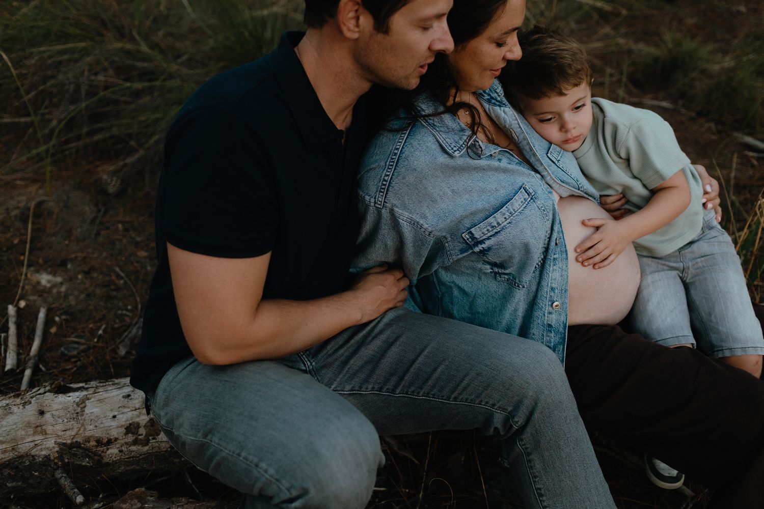 Smiling father and pregnant mother sit together outdoors with their young son leaning against her baby bump.