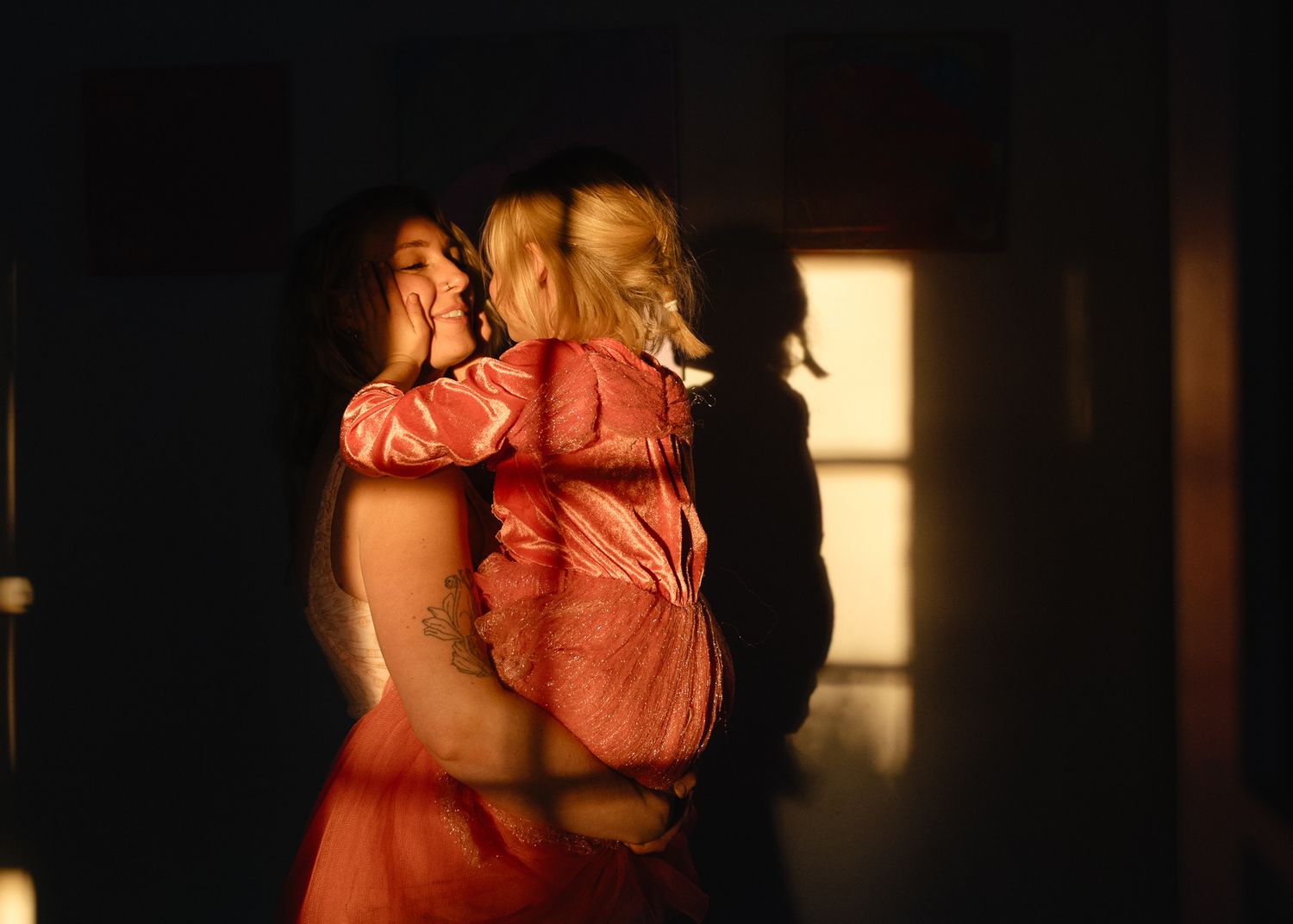 A mother in a red dress holds her young blonde daughter in warm golden light beside a glowing window.