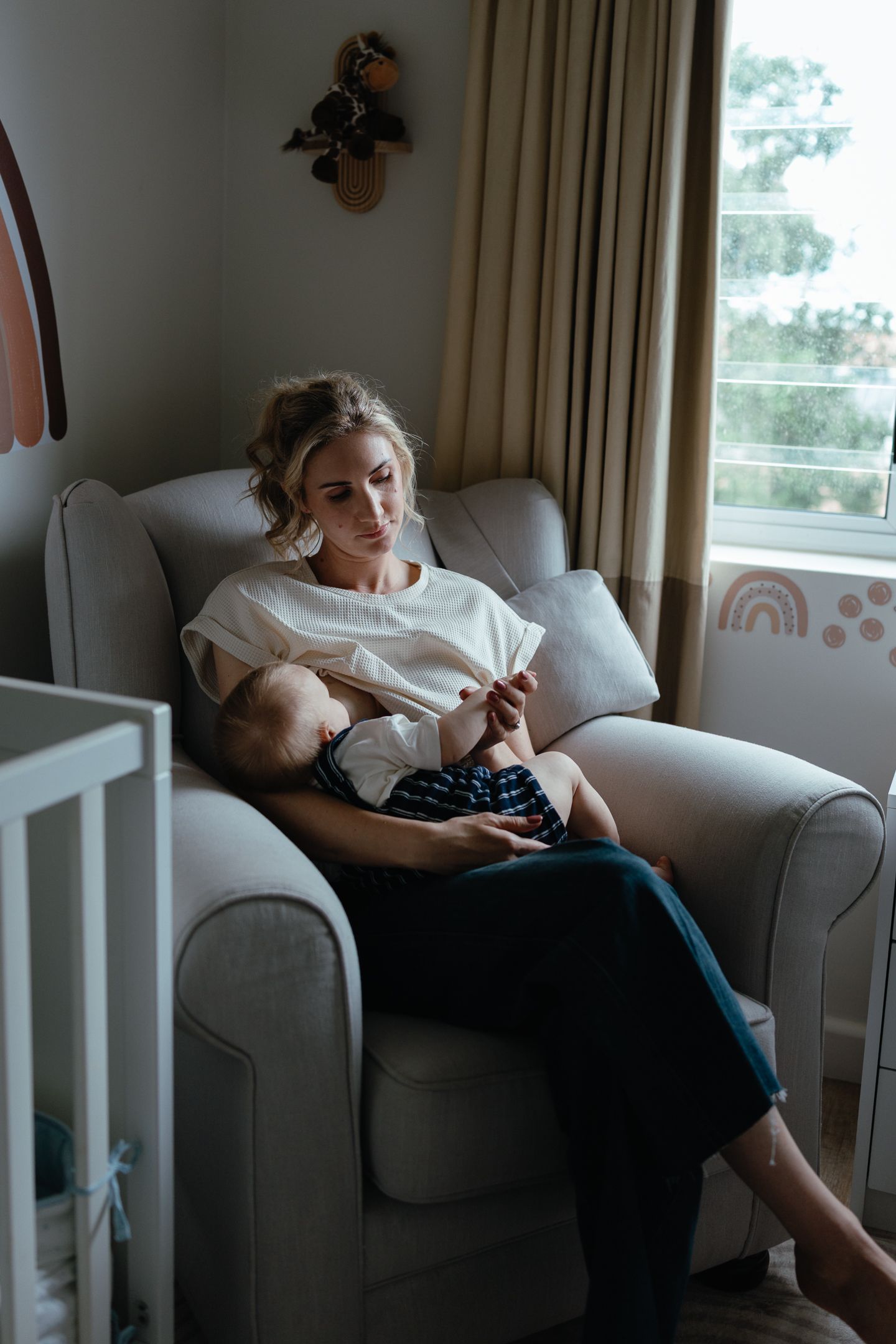 Mother in a cream top sits in a gray armchair by a window, gazing down at her baby in a sunlit nursery.