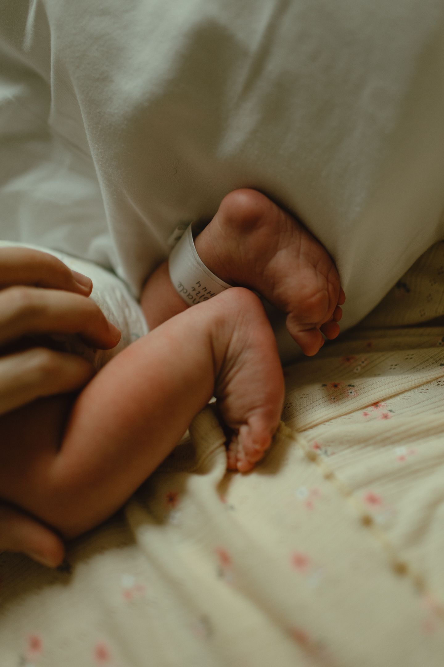 Newborn baby's tiny feet with a hospital ID bracelet rest on a floral bedsheet beside a parent's hand.