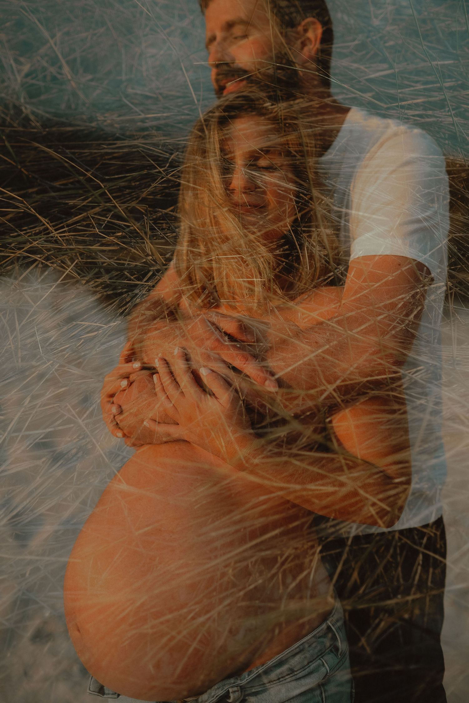 A bearded man in a white t-shirt embraces a pregnant woman from behind, framed by tall dune grass in warm light.