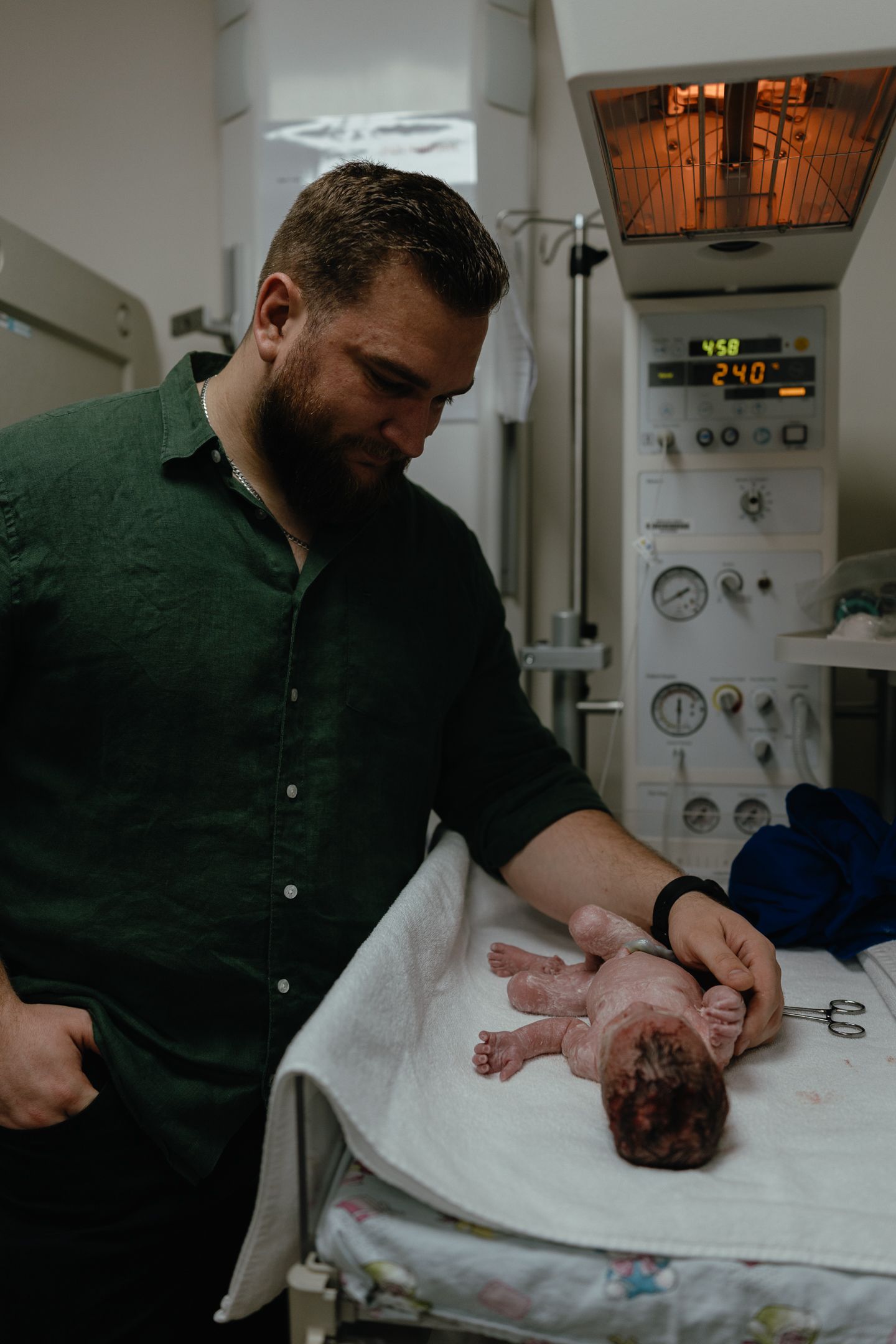 A bearded man in a green shirt gently touches a newborn baby lying on a warmed hospital bassinet under a radiant heater.