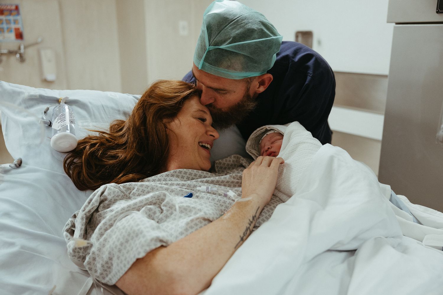 Smiling mother in a hospital gown cradles her newborn while the father in scrubs kisses her head in the delivery room.