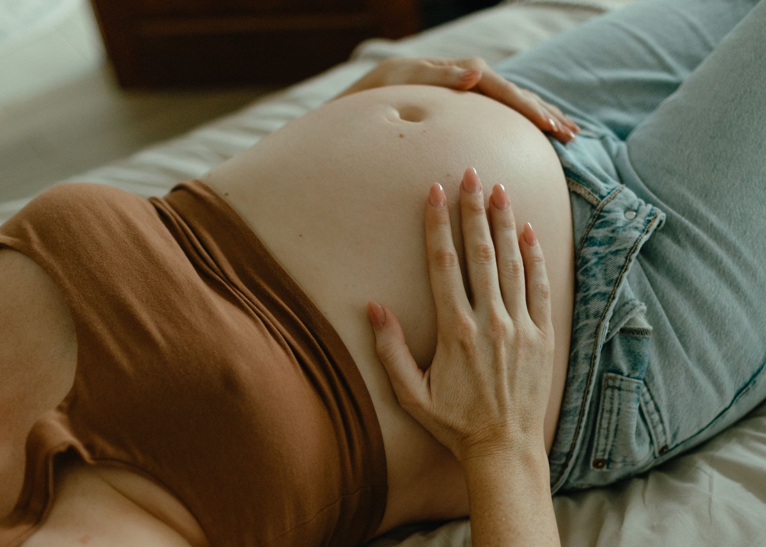 Close-up of a pregnant woman in a brown top and jeans resting her hands gently on her bare belly.