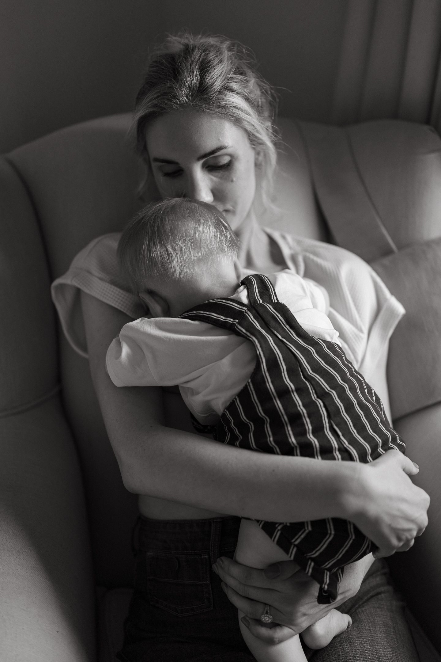A black and white photo of a mother in a loose top cuddling a baby in a striped outfit on a cozy armchair.
