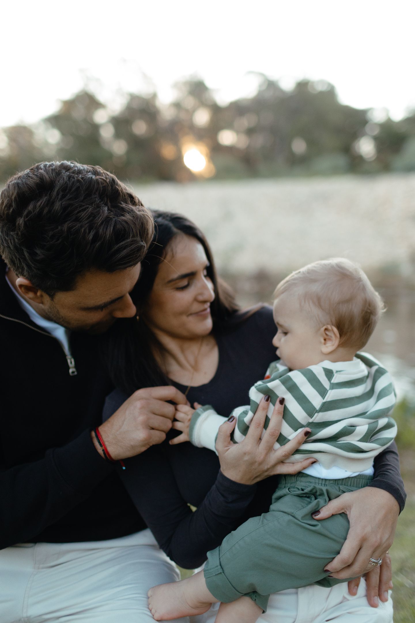 A smiling mother and father hold their baby boy in a striped sweater during a cozy outdoor family portrait at sunset.
