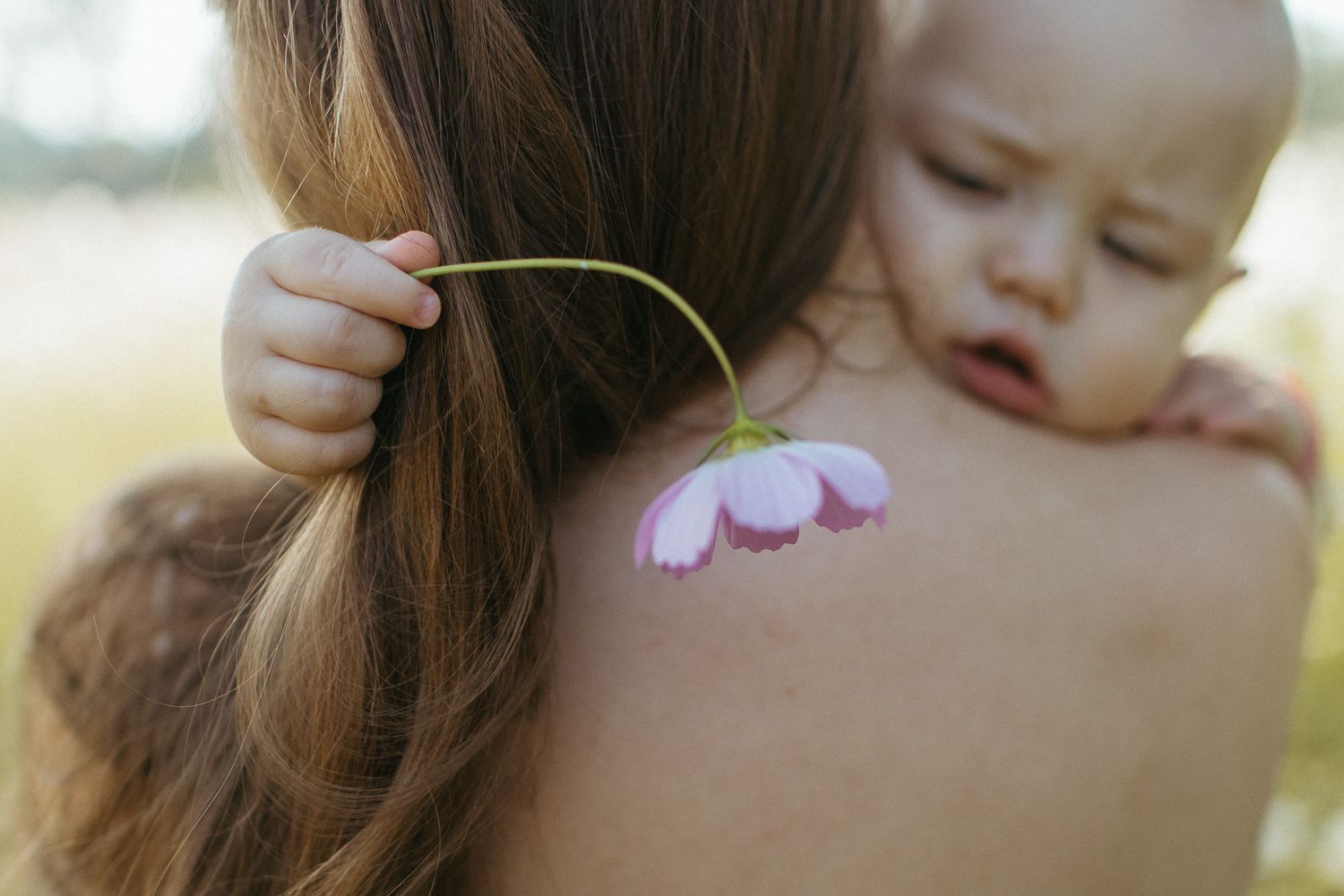 Sleepy baby rests on a mother's bare shoulder while holding a delicate pink flower near her long brown hair.
